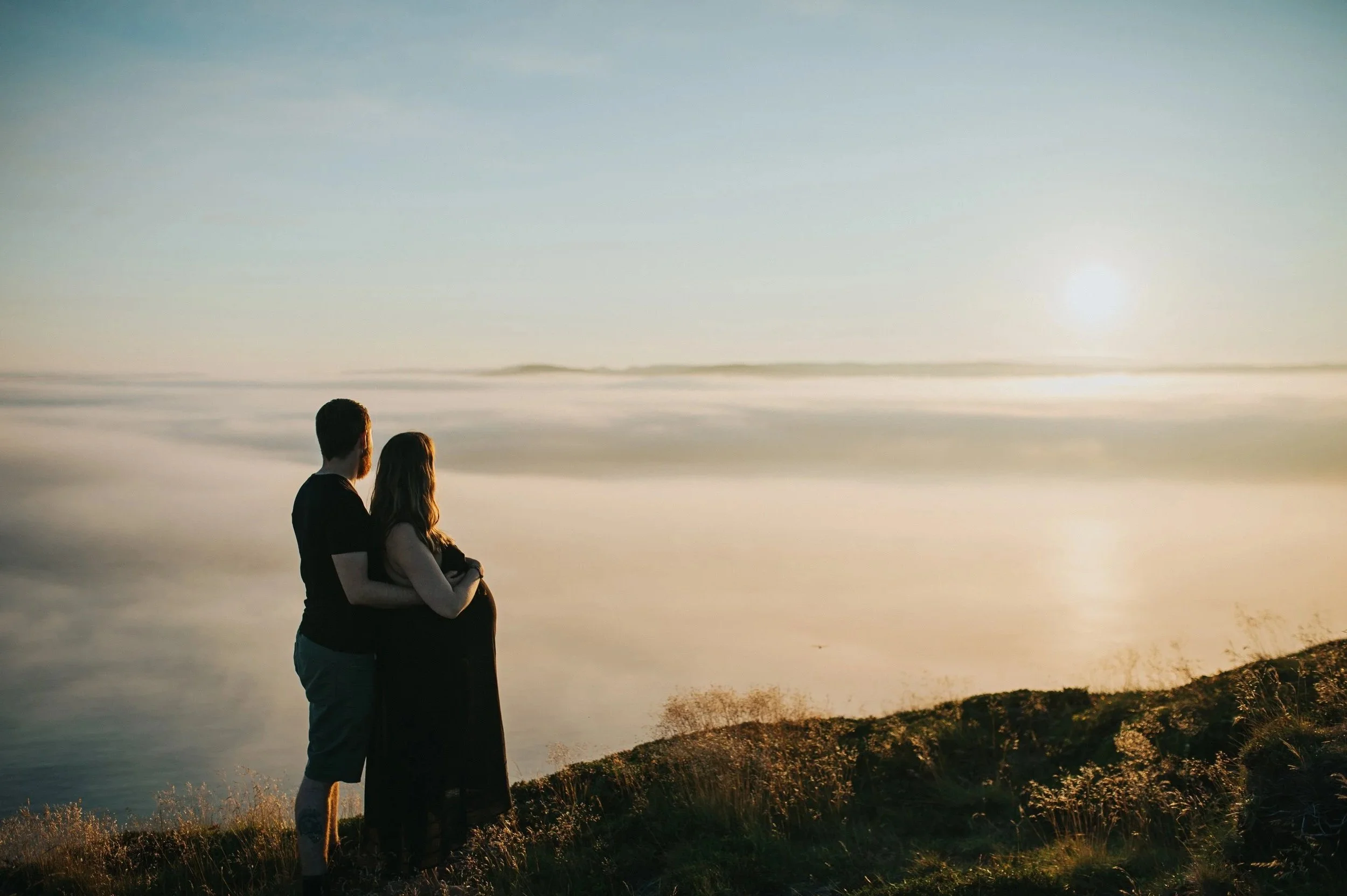 A couple stands together on a grassy hill at sunset, overlooking a large body of water with distant land on the horizon.