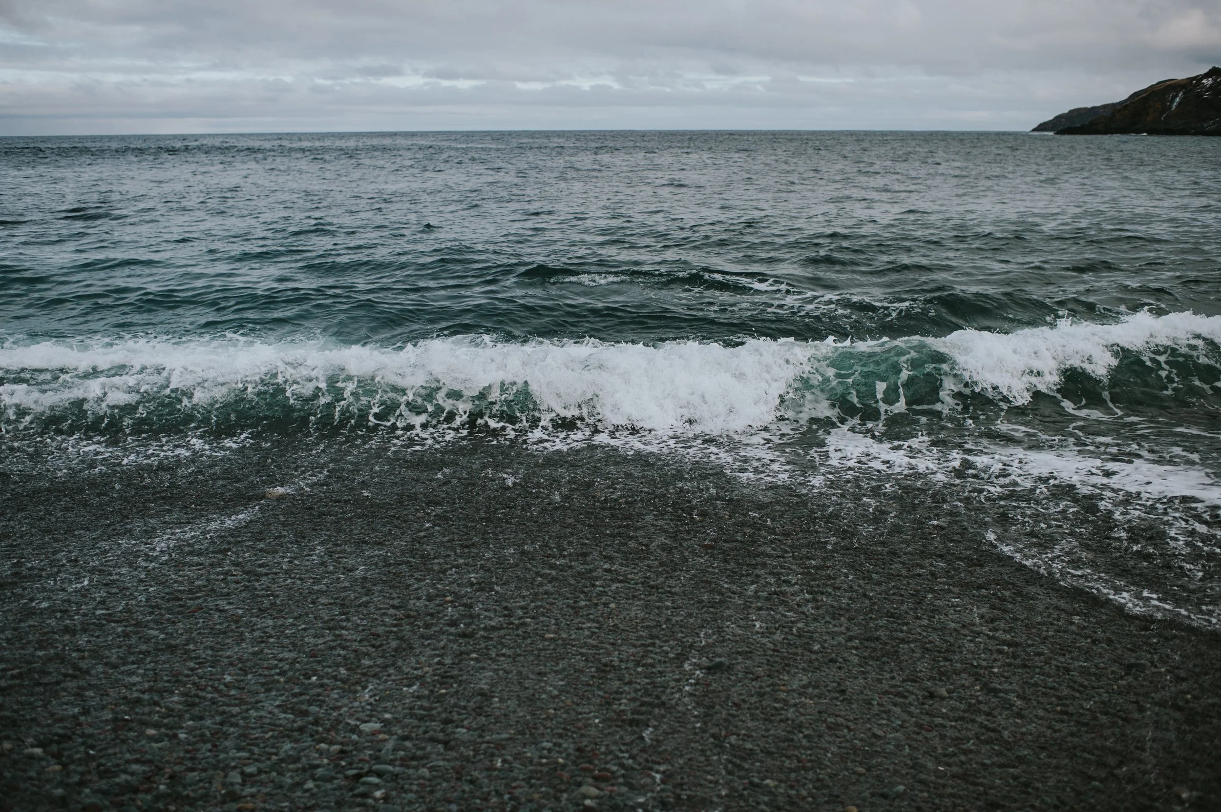 Ocean waves washing onto a rocky shore with a cloudy sky and distant hills.