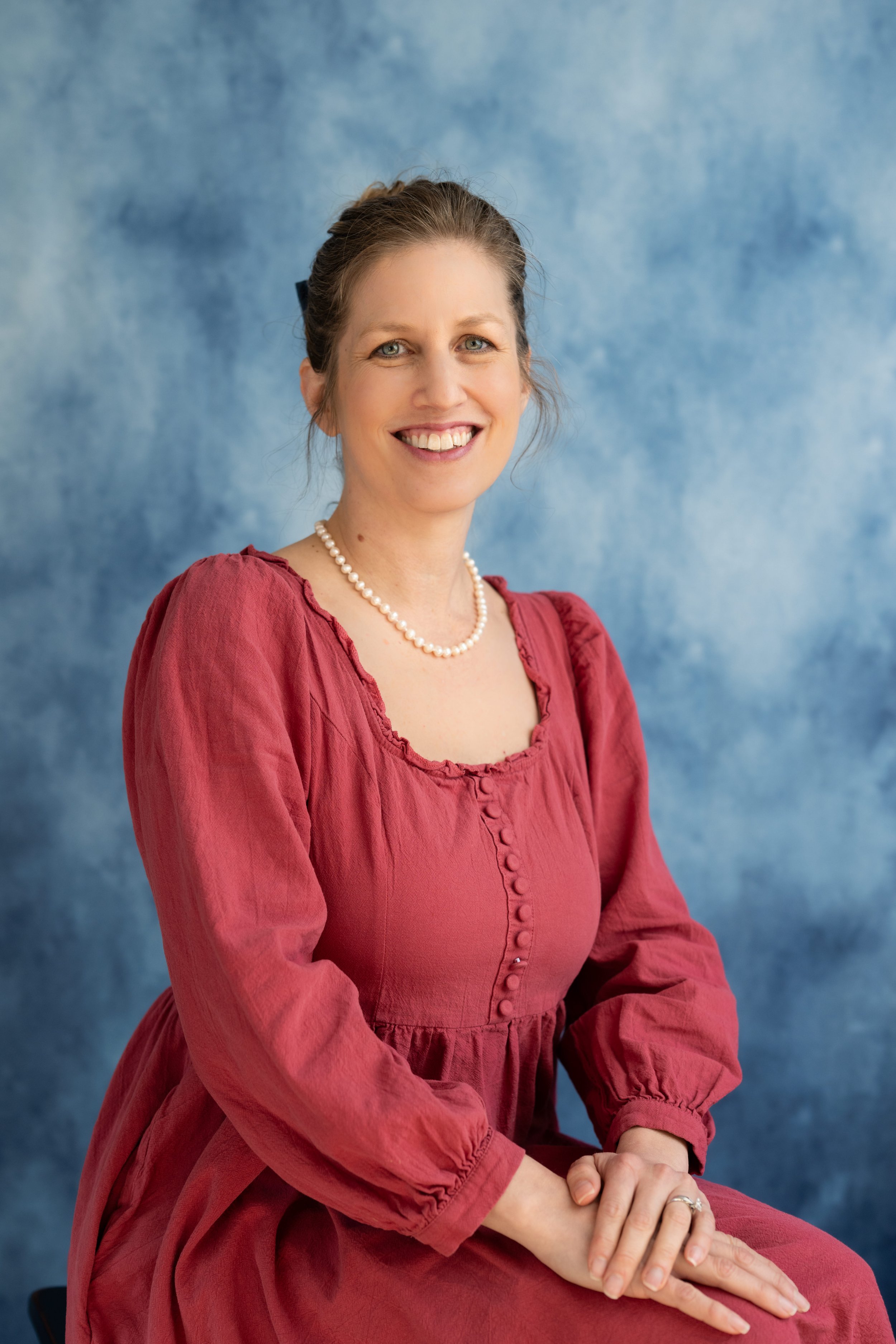 A woman with brown hair pulled back, wearing a red dress with a pearl necklace, smiling with hands clasped, seated against a blue textured background.