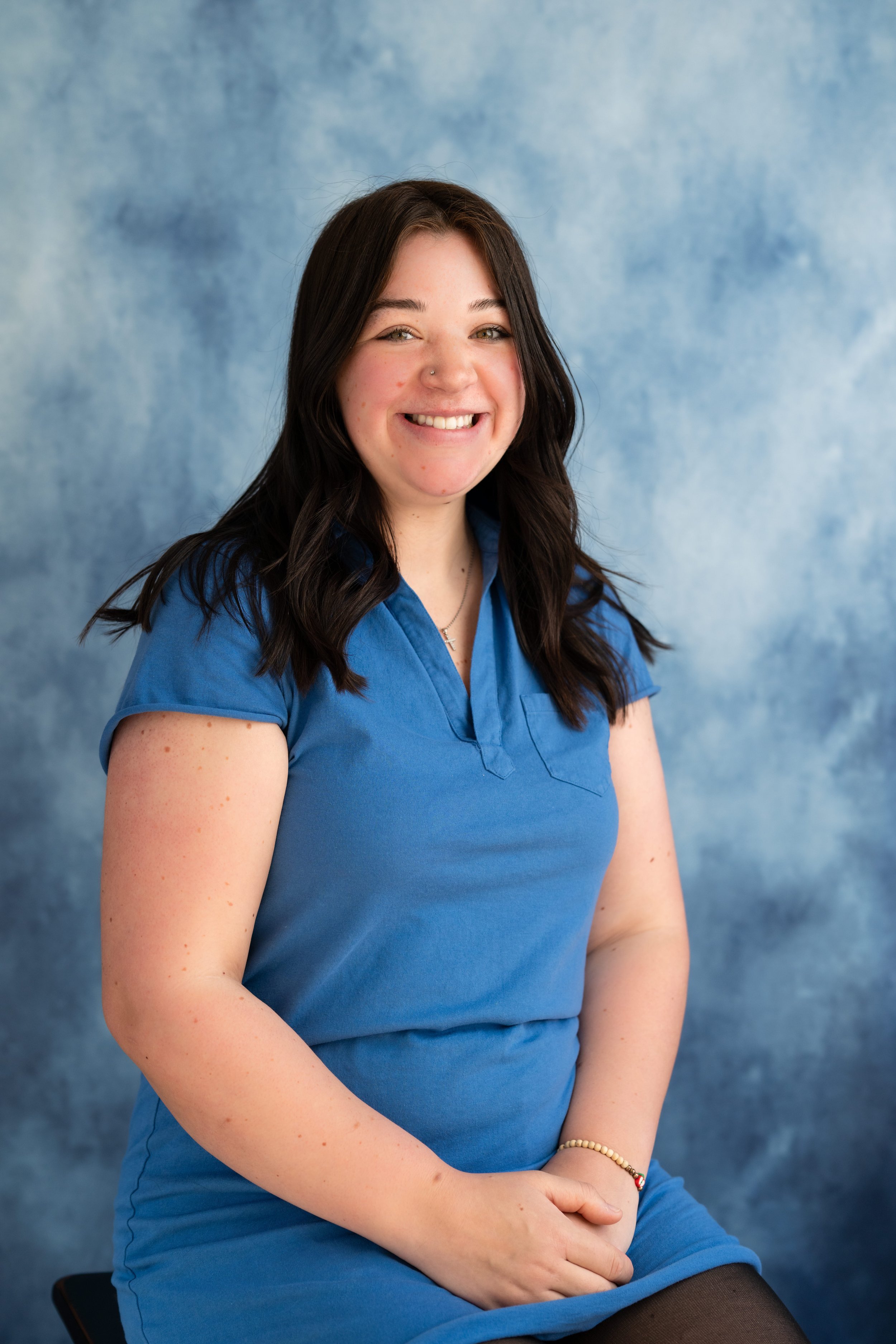 Portrait of a young woman with long dark hair, smiling, wearing a blue shirt and black stockings, sitting against a mottled blue background.