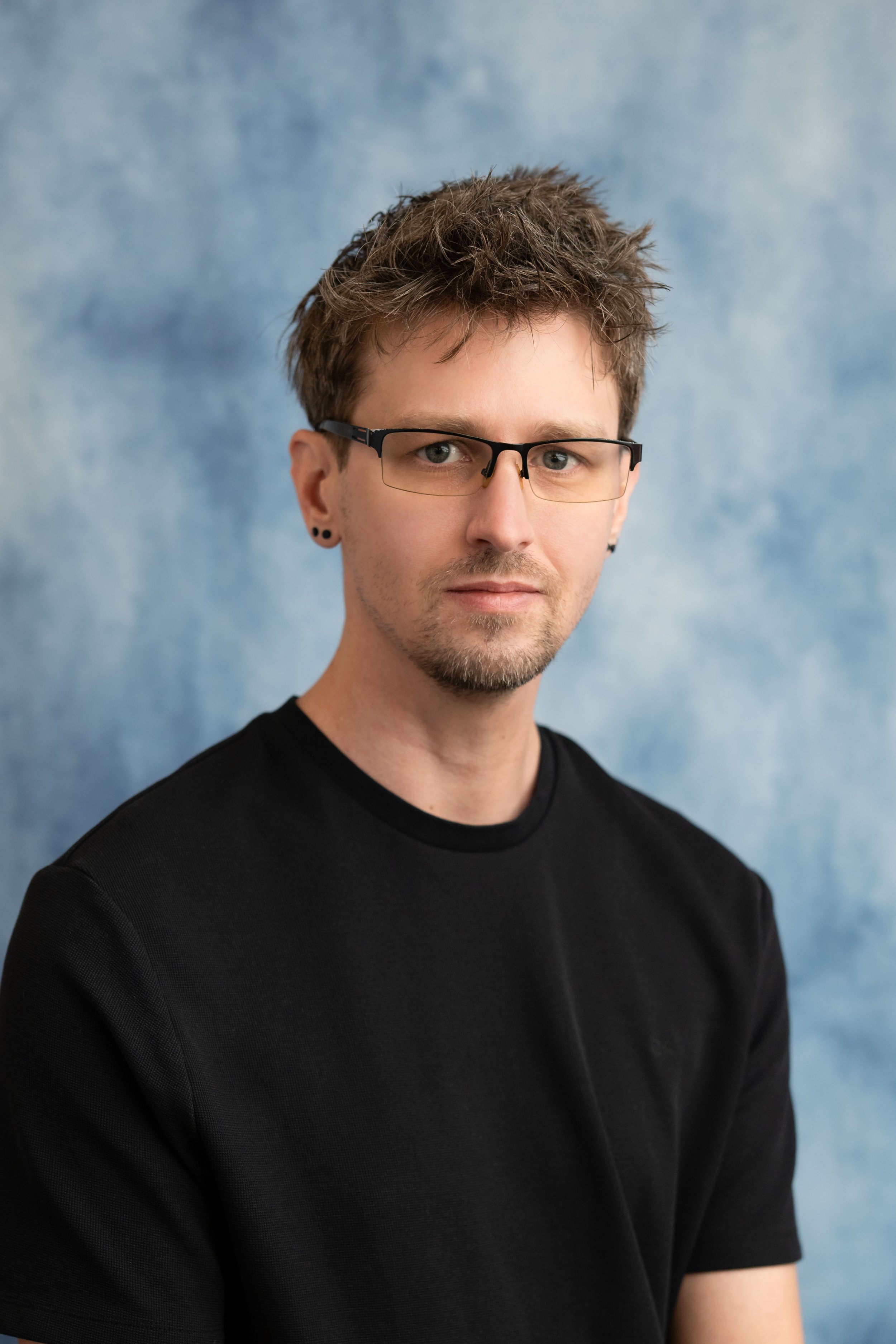A young man with short, spiky brown hair, wearing glasses and black earrings, dressed in a black shirt, standing against a blue textured background.