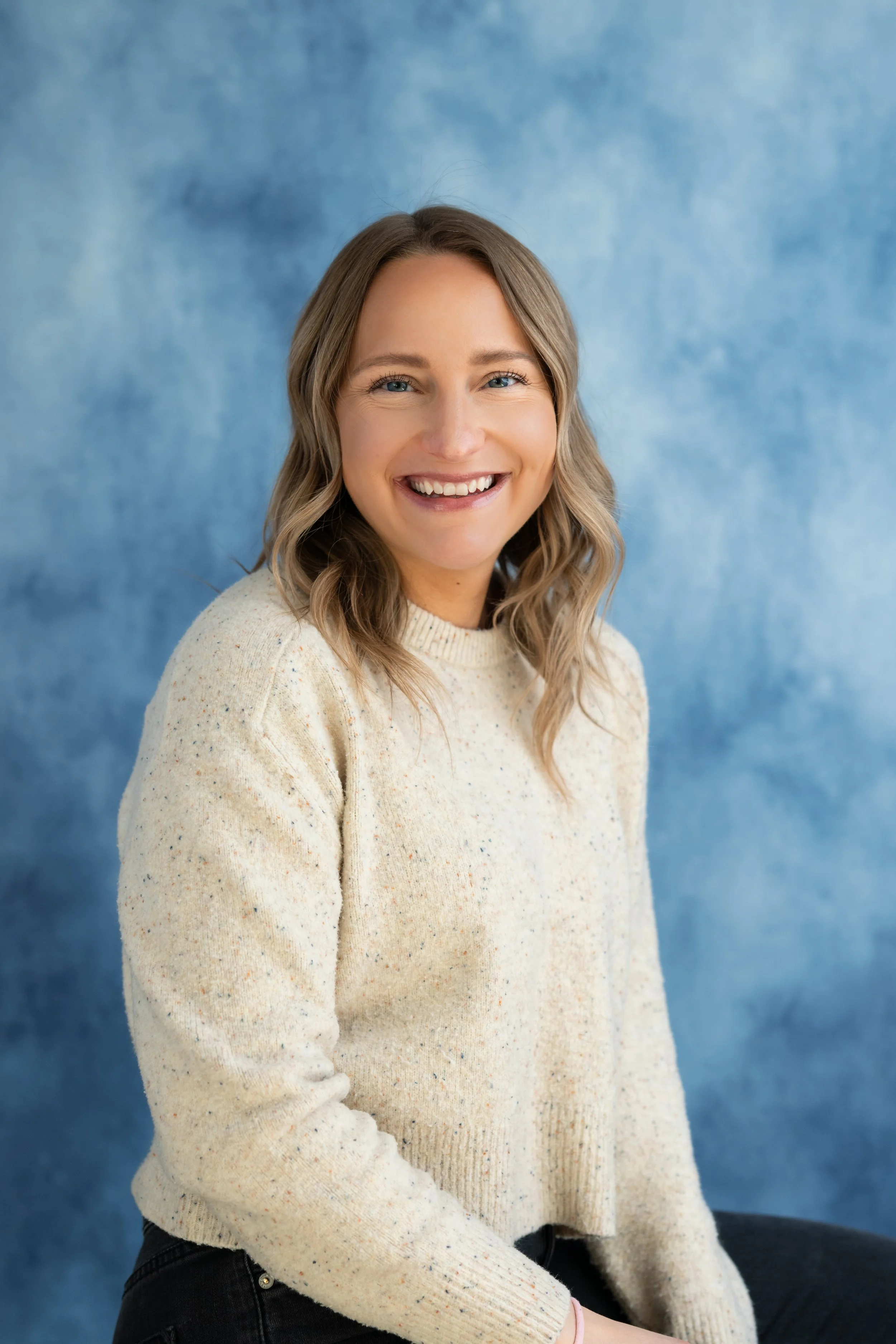 A young woman with wavy blonde hair smiling, wearing a cream-colored speckled sweater, seated against a blue textured background.