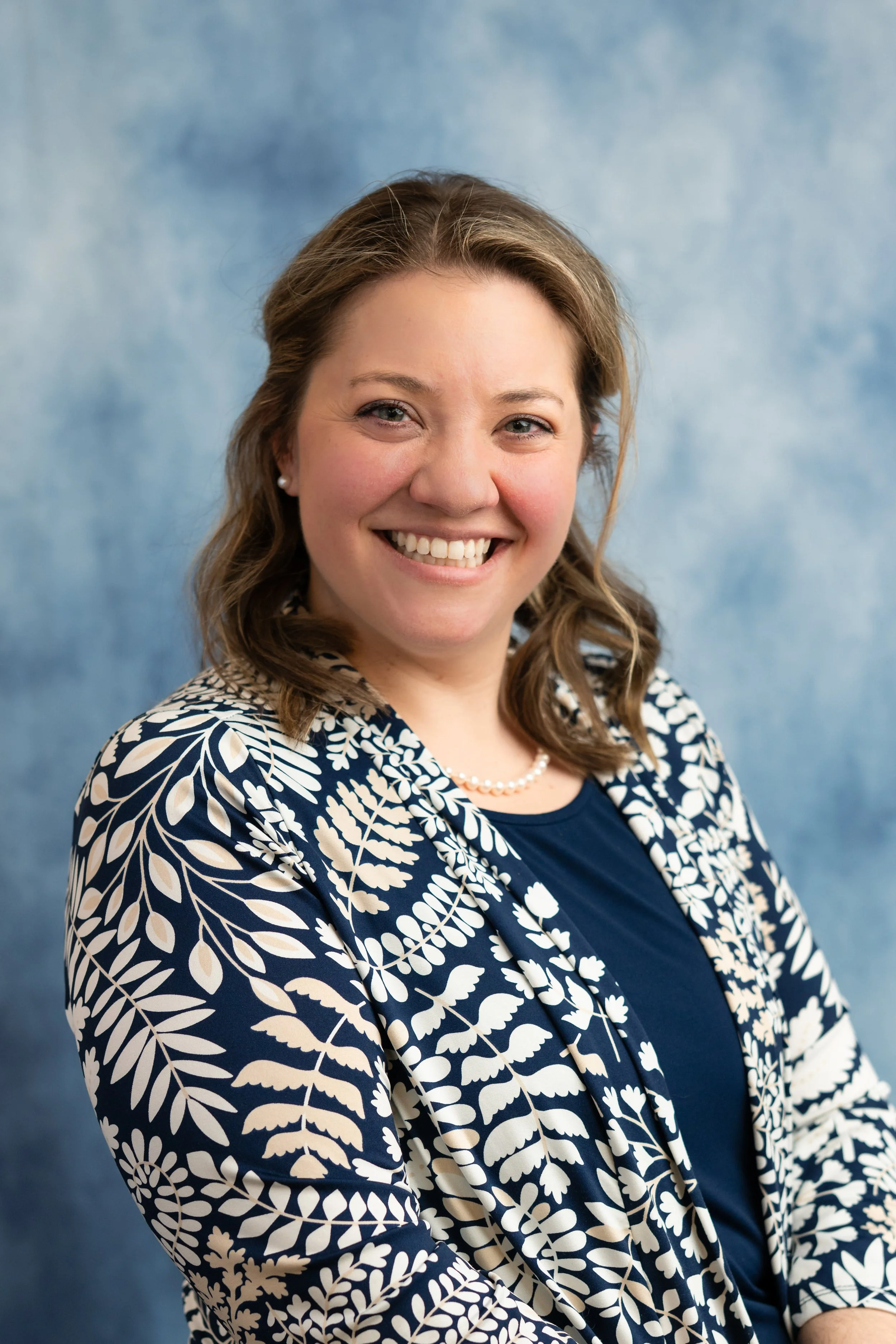 A woman with light brown hair, wearing pearl earrings, a pearl necklace, a navy blue top, and a navy and white patterned blazer, smiling in front of a blue textured background.