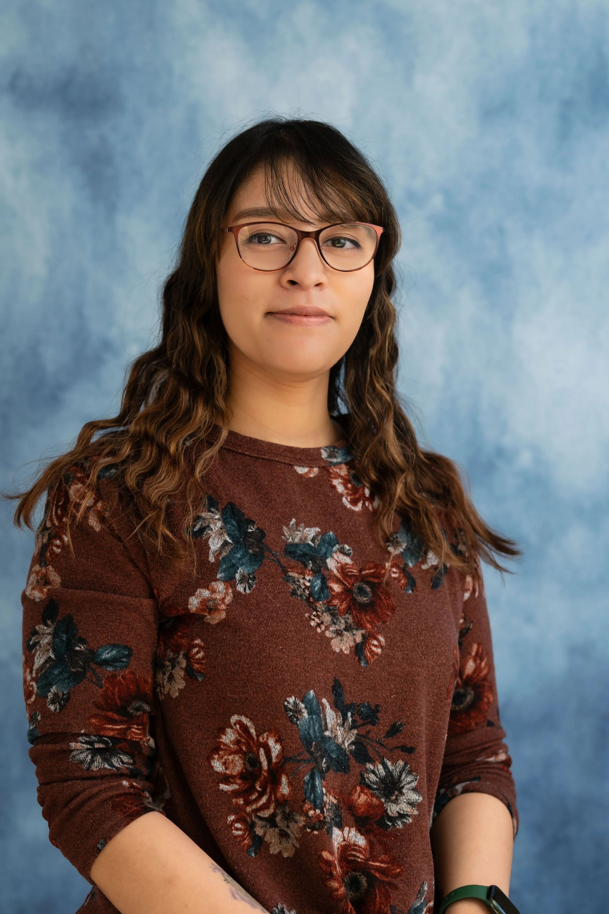 A woman with glasses and wavy brown hair, wearing a maroon floral sweater, stands against a blue textured background.