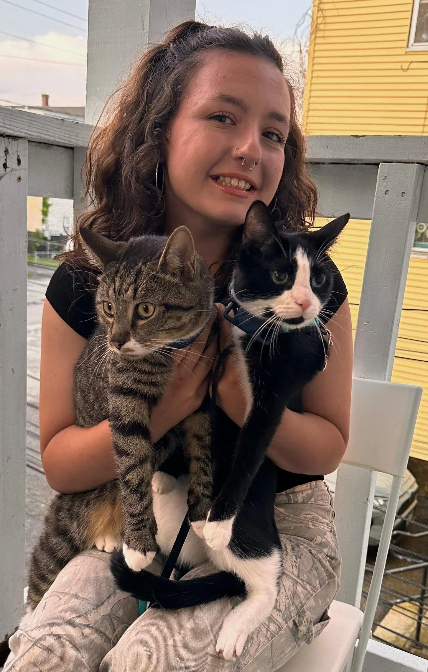 A young woman with brown curly hair and a nose piercing smiling and holding two cats on her lap, sitting outdoors near a yellow house and a wooden railing.