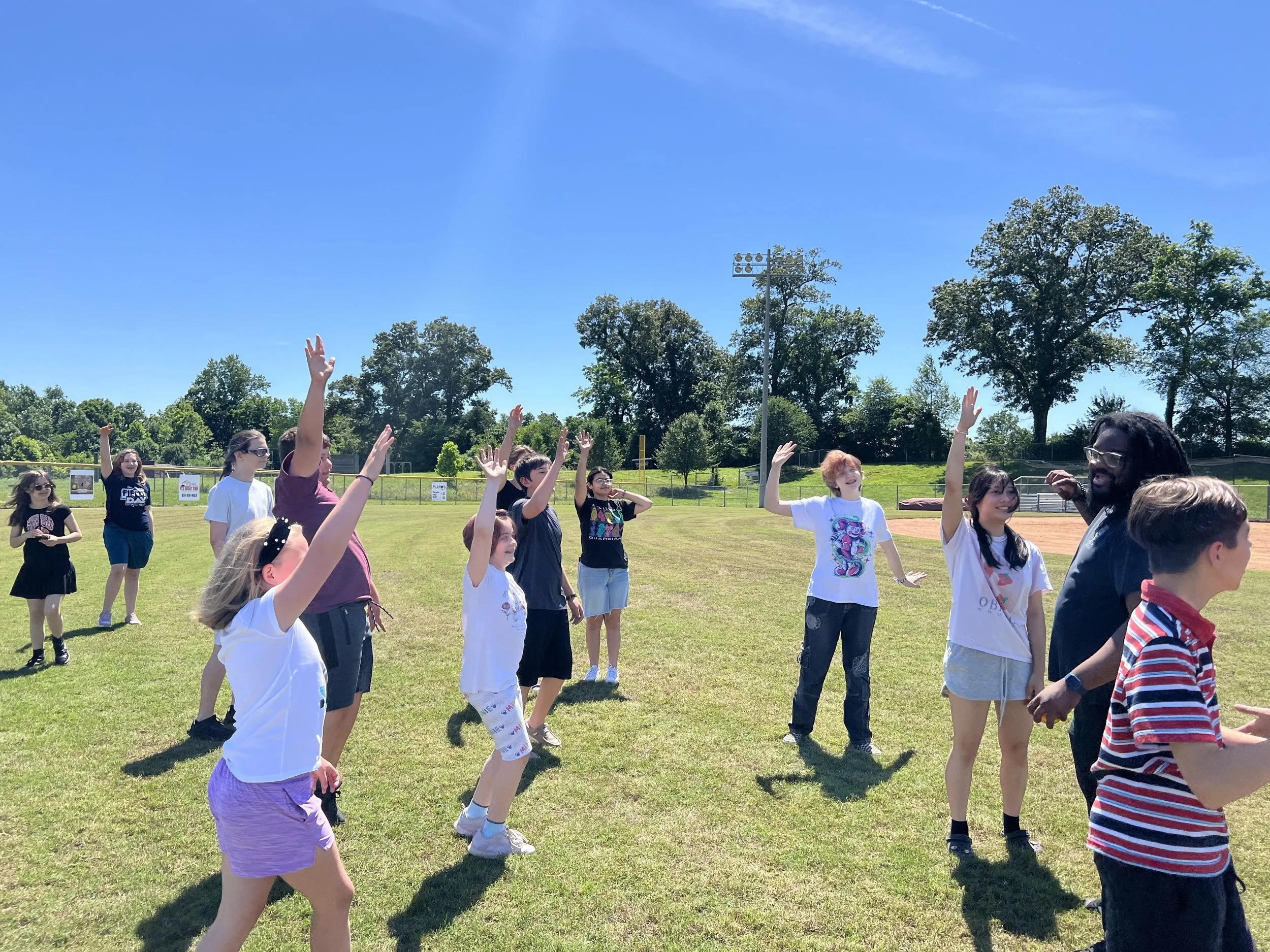 Children and an adult on a sunny grassy field, engaging in an outdoor activity, with some raising their hands, others smiling, and trees and a clear blue sky in the background.