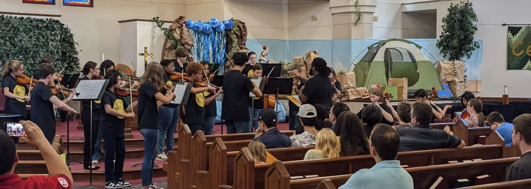 Youth orchestra performing on stage inside a church or community hall, with audience seated on benches. Stage decorated with camping tent, cardboard boxes, and a backdrop resembling a forest or outdoor scene.