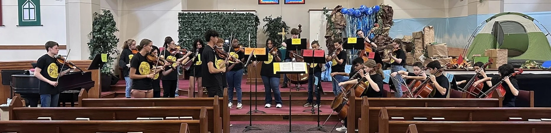 A group of children playing violins, cellos, and a pianist performing on stage inside a church or auditorium with wooden pews, decorated with Christmas displays in the background.