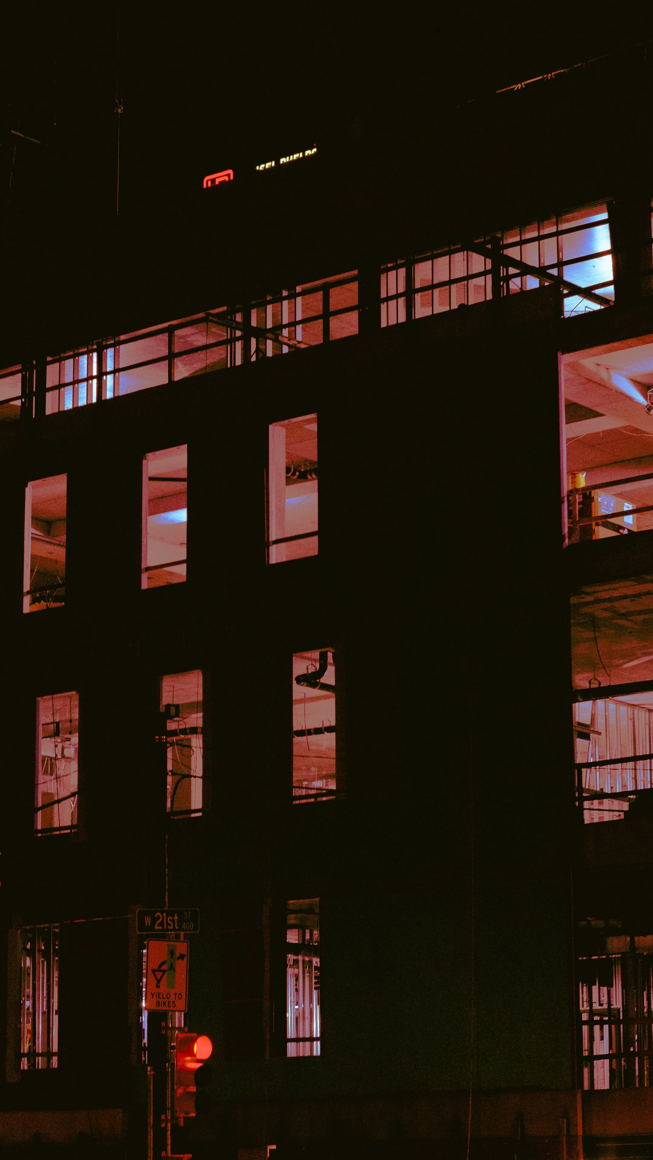 Nighttime view of a modern multi-story building with illuminated open interior spaces, visible through large windows, and a street sign at the bottom left corner reading 'W 21st St'.