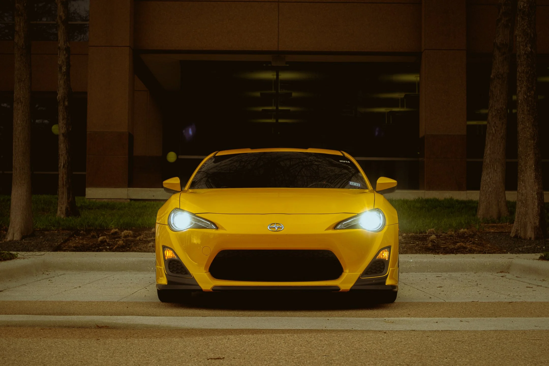 A yellow sports car is parked on a city street at night, with trees and a modern building in the background. Automotive Photos in Dallas Texas