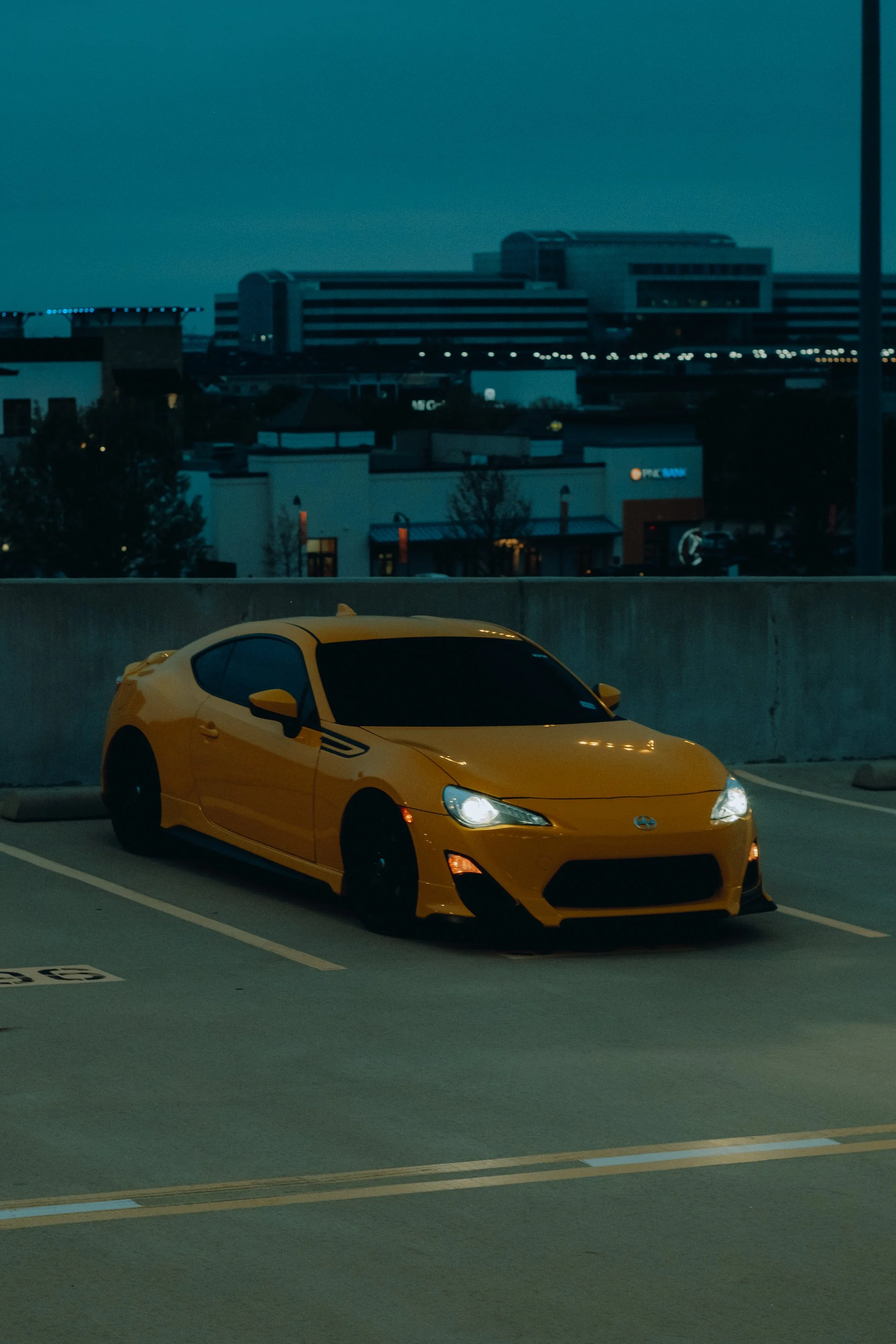 Yellow sports car parked in a parking lot at dusk with city buildings in the background. Taken in Dallas Texas