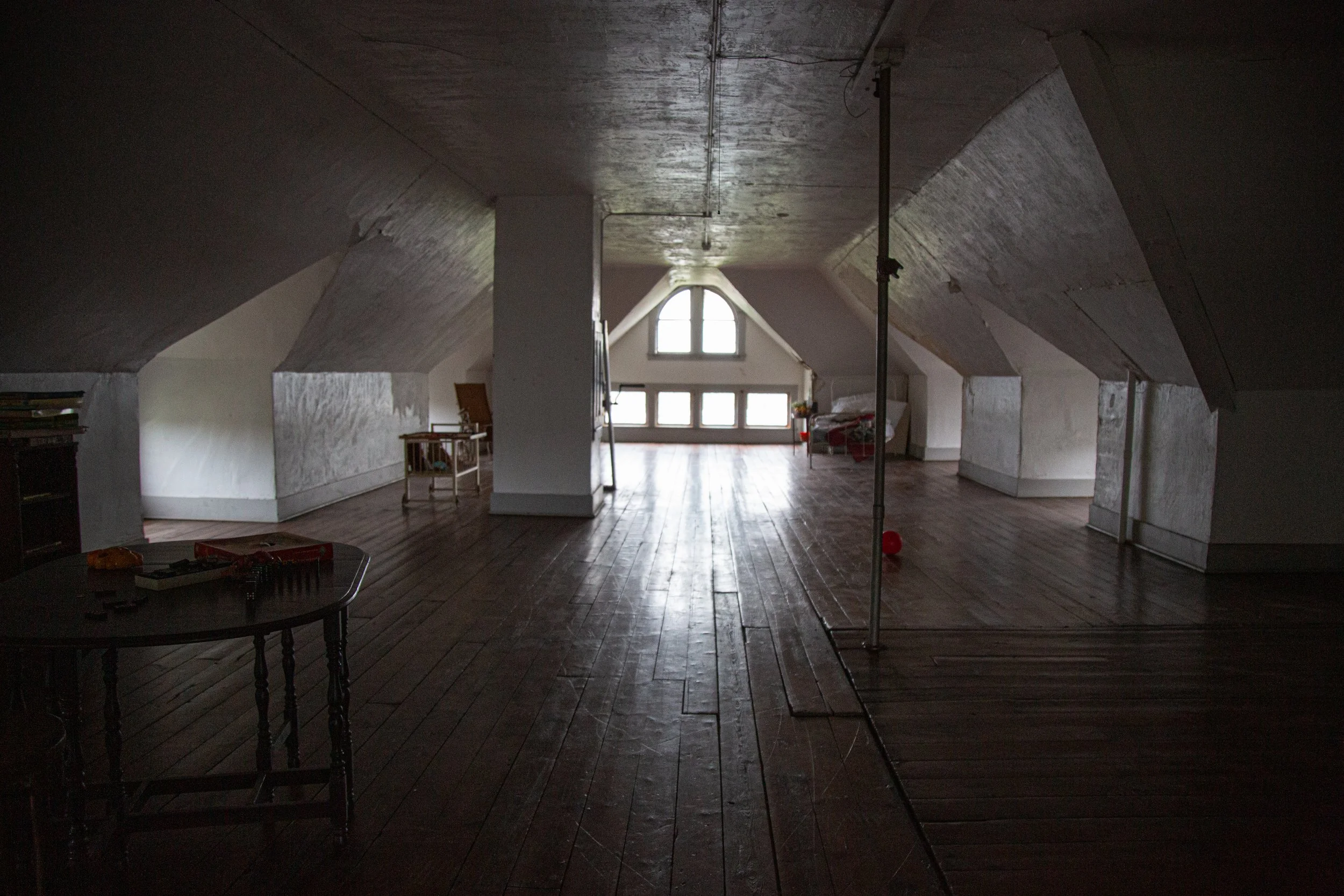 An attic room with wooden floors, a wheelchair near a window, a green chair, and a crutch leaning against the wall, with peeling paint and a damaged ceiling.