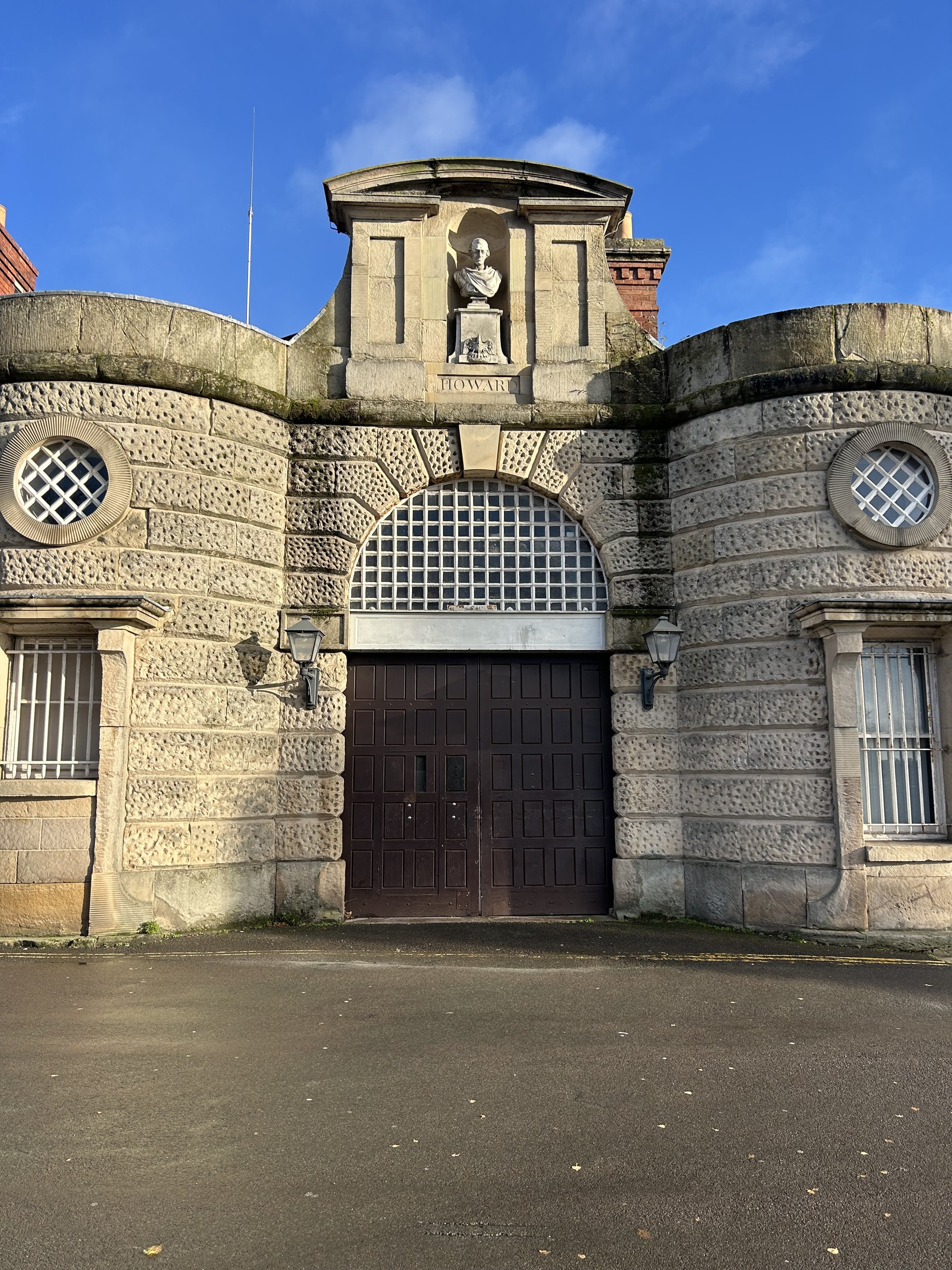 Historic stone building with a large wooden gate, decorative circular windows, two wall-mounted lanterns, and a bust sculpture of a man above the gate with the name 'Howard' beneath it. Clear blue sky in the background.