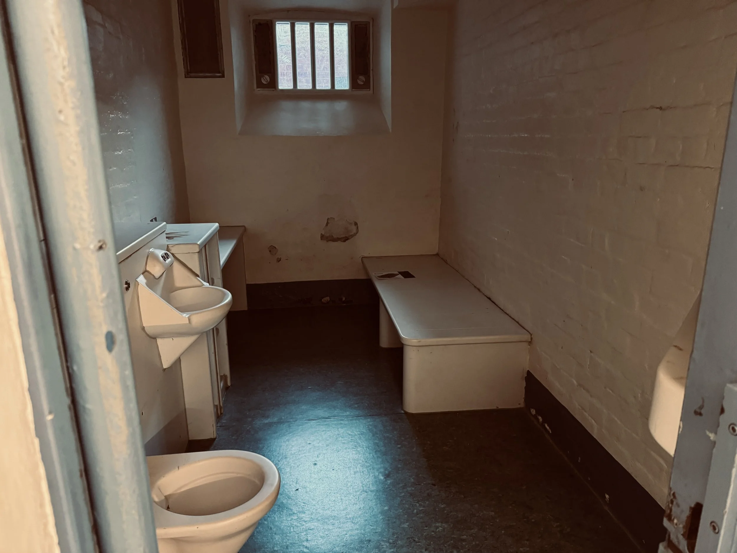 A small, dimly lit room with bare brick and plaster walls, a toilet in the foreground, a small sink with a cabinet next to it, a window on the back wall letting in some light, and a bench or table along the right wall.