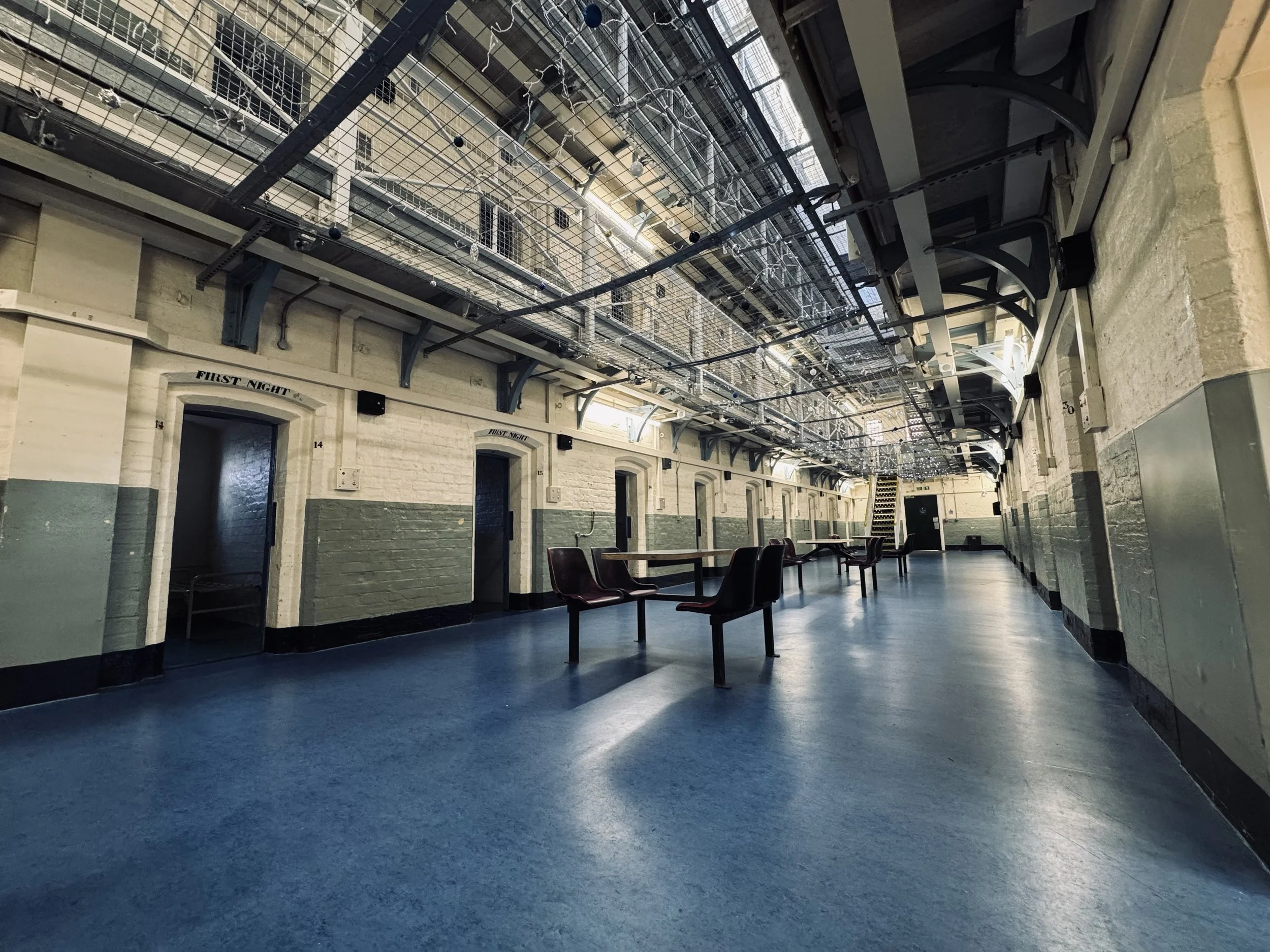 Interior of a historic prison with high ceilings, brick walls painted white and green, and several prison cell doors. There are some chairs and tables in the corridor, and an overhead steel walk in the upper part of the building.