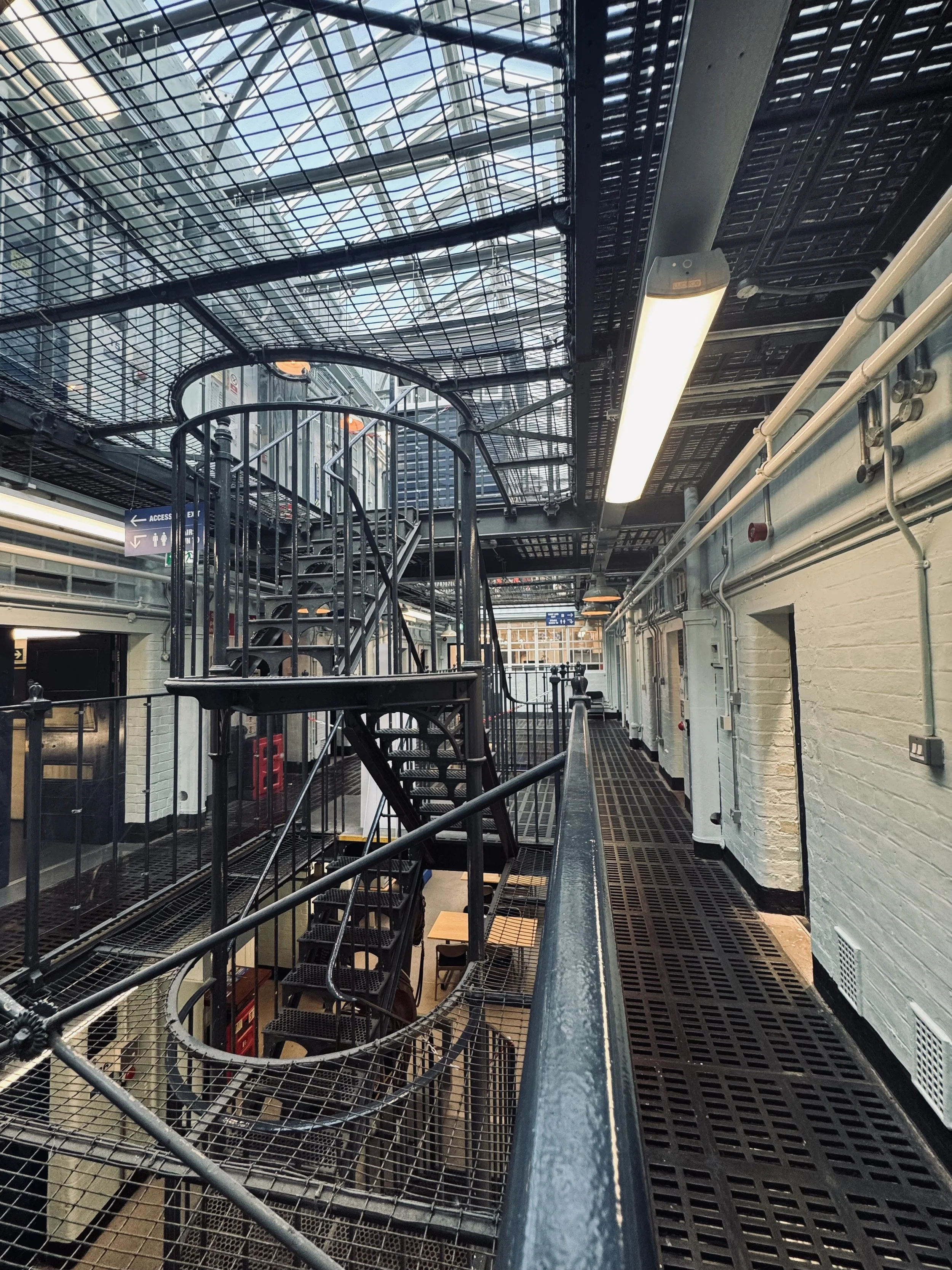 Industrial-style interior with metal staircase, metal grating floors, white brick walls, and exposed piping, illuminated by ceiling lights.