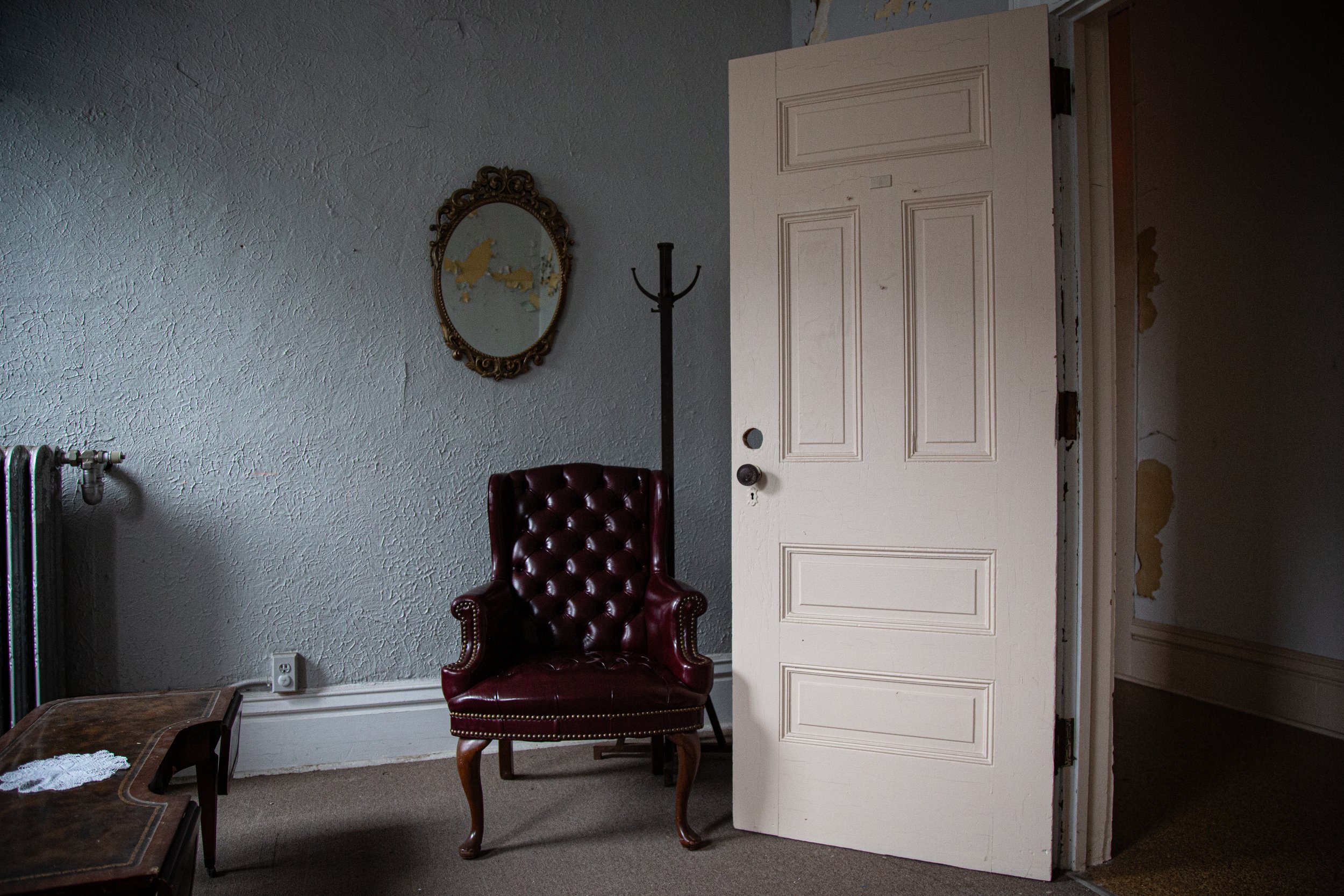 A dimly lit room with an old patterned green wall, a wooden rocking chair, a vintage baby carriage with a striped blue interior, a red wooden cradle, and a window with peeling paint revealing a brick building outside.