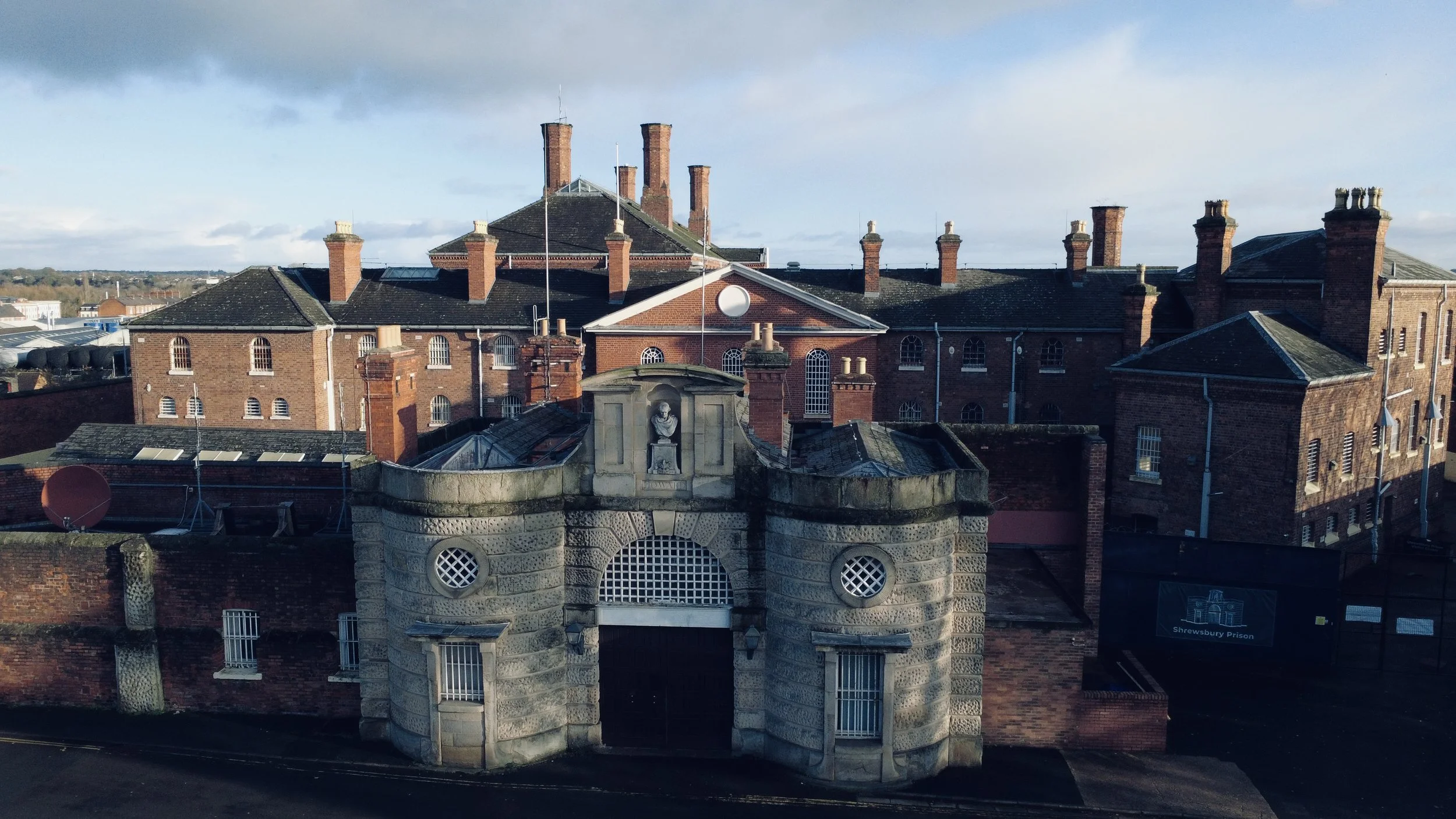 A historic prison building with a stone entrance gate, a sculpture above it, and multiple brick buildings with chimneys in the background, marked as Shrewsbury Prison.