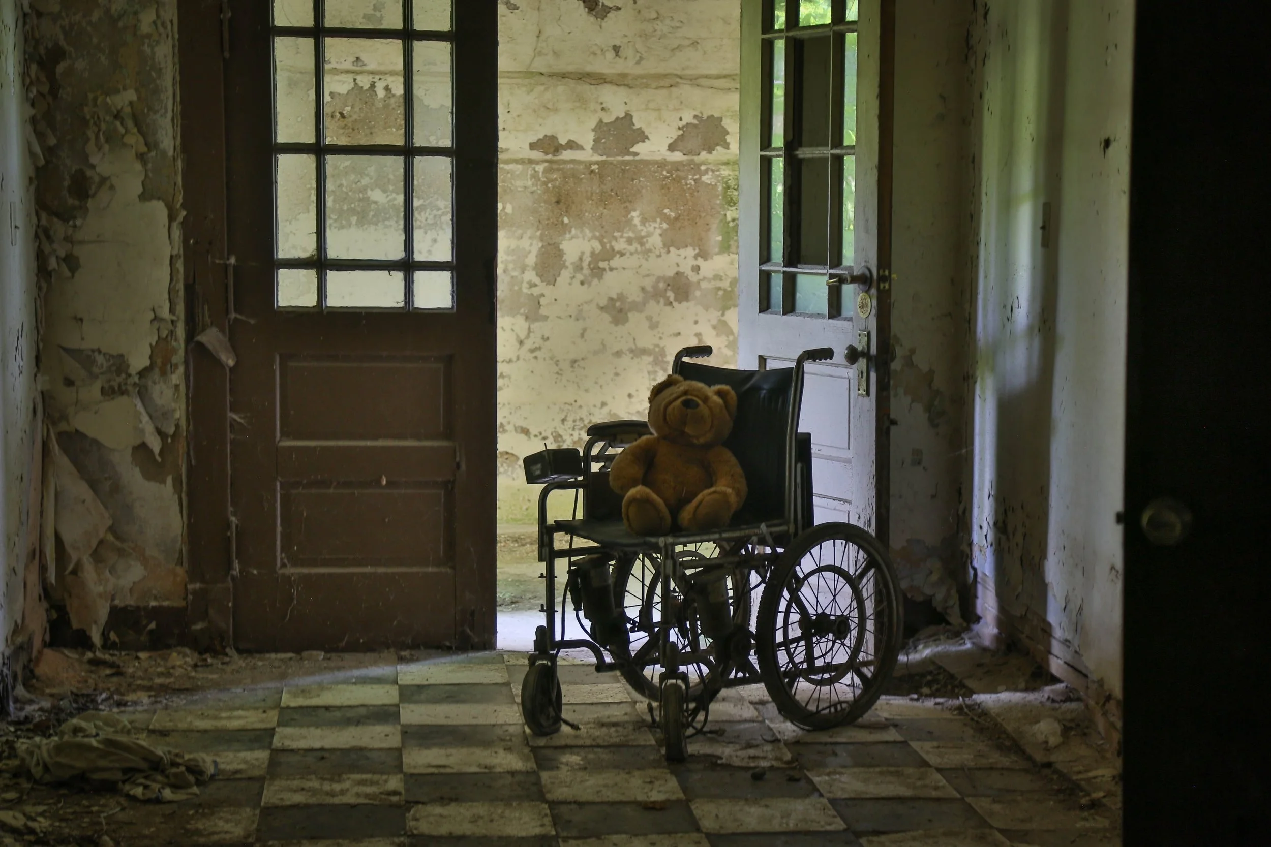 Silhouette of a wheelchair inside a dark room with a partially open door revealing a weathered wall outside.