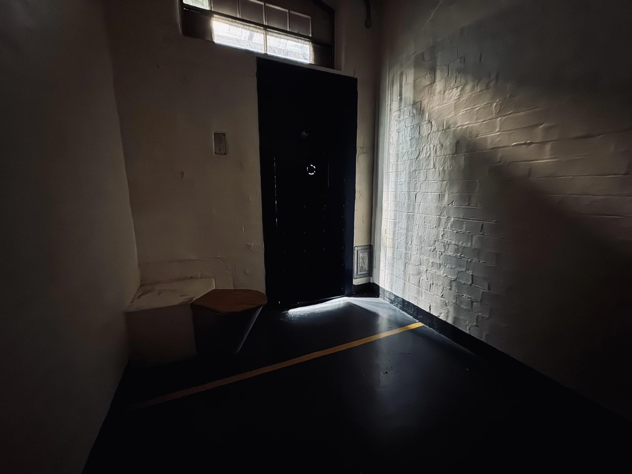 Dimly lit interior of a room with a black door slightly open, allowing some light to spill in. White brick walls and a small window near the ceiling are visible. A white toilet with a wooden seat is in the corner, and the floor has a yellow line running along the edge.
