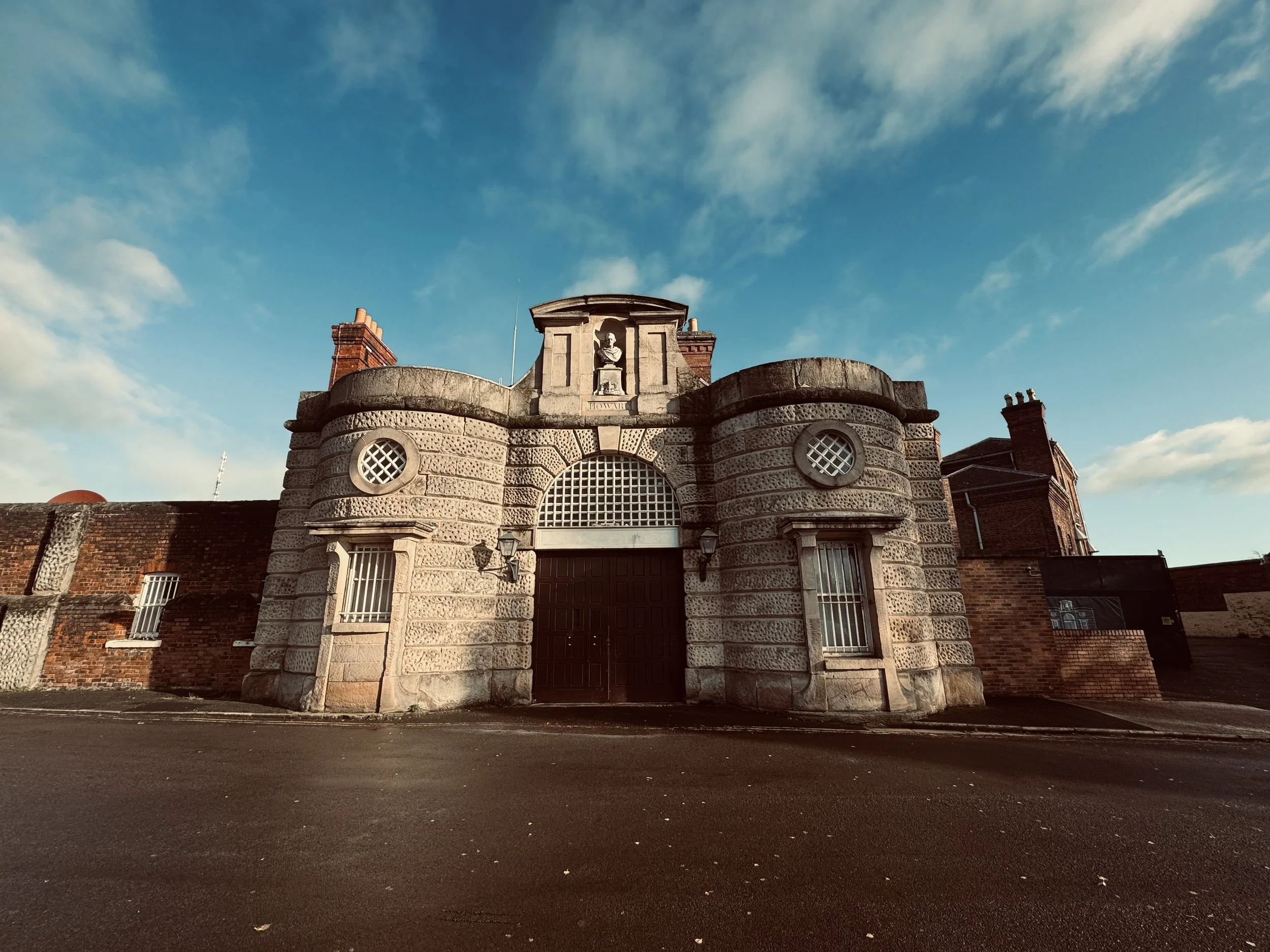 Front view of a historic building with stone and brick architecture, arched entrance, small windows with bars, and a statue above the entrance, under a blue sky with clouds.