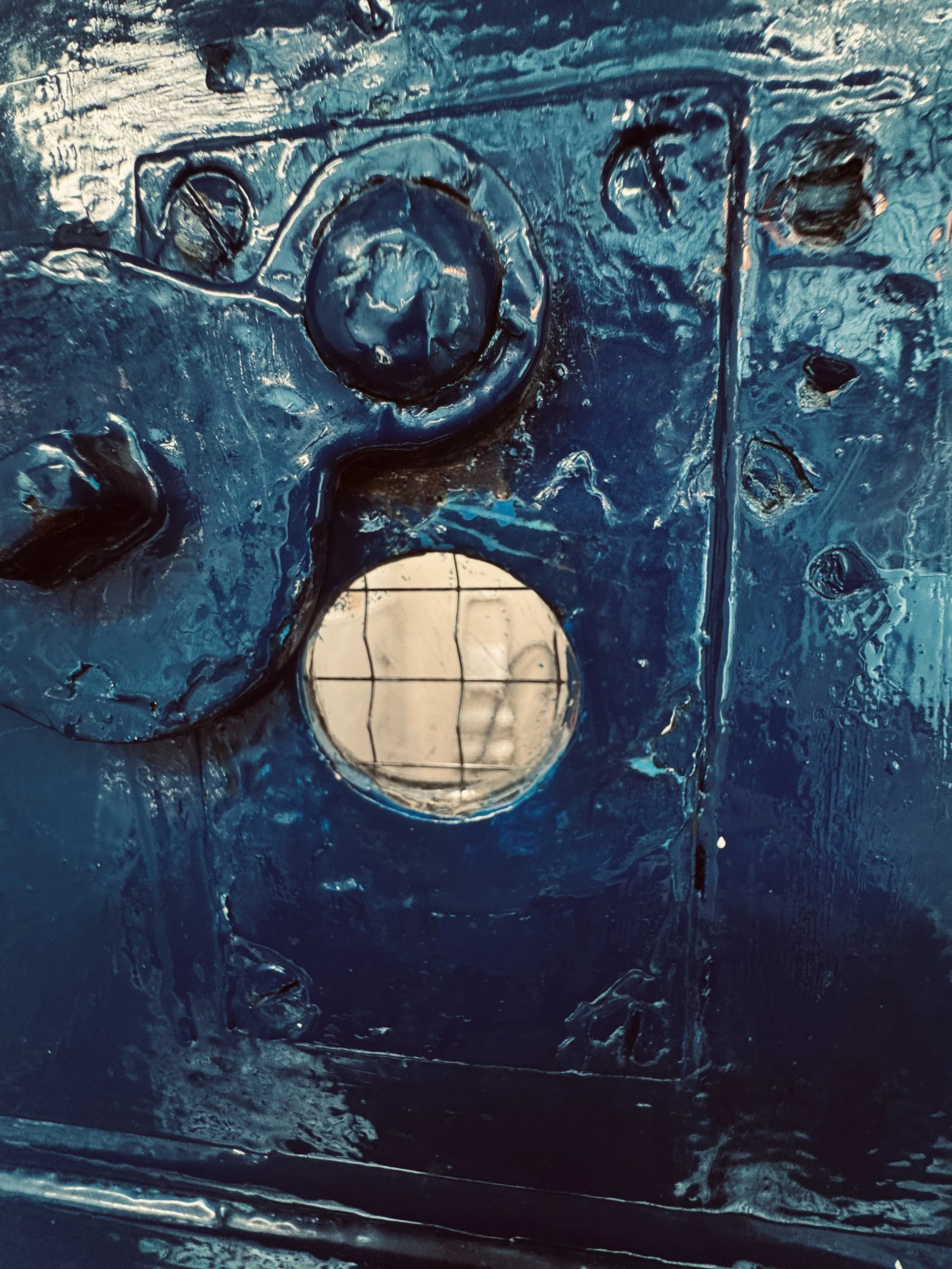 Close-up of a metallic blue painted part with a round hole showing a wire grid behind it, surrounded by several bolts and screws.