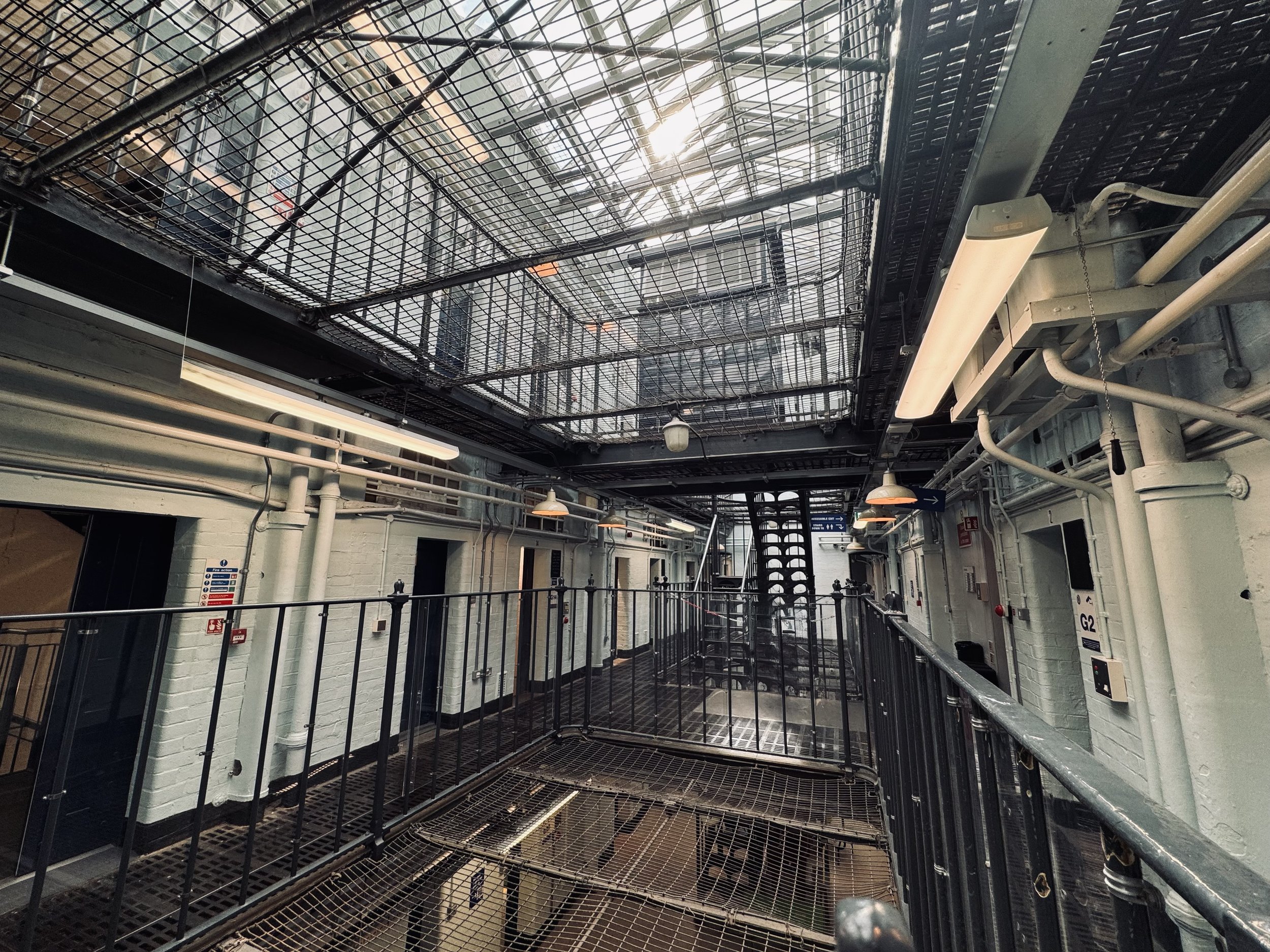 Interior view of a multi-level industrial or modern building with metal walkways, railings, lattice ceiling, and a staircase at the center, with some lighting fixtures and piping along the walls.