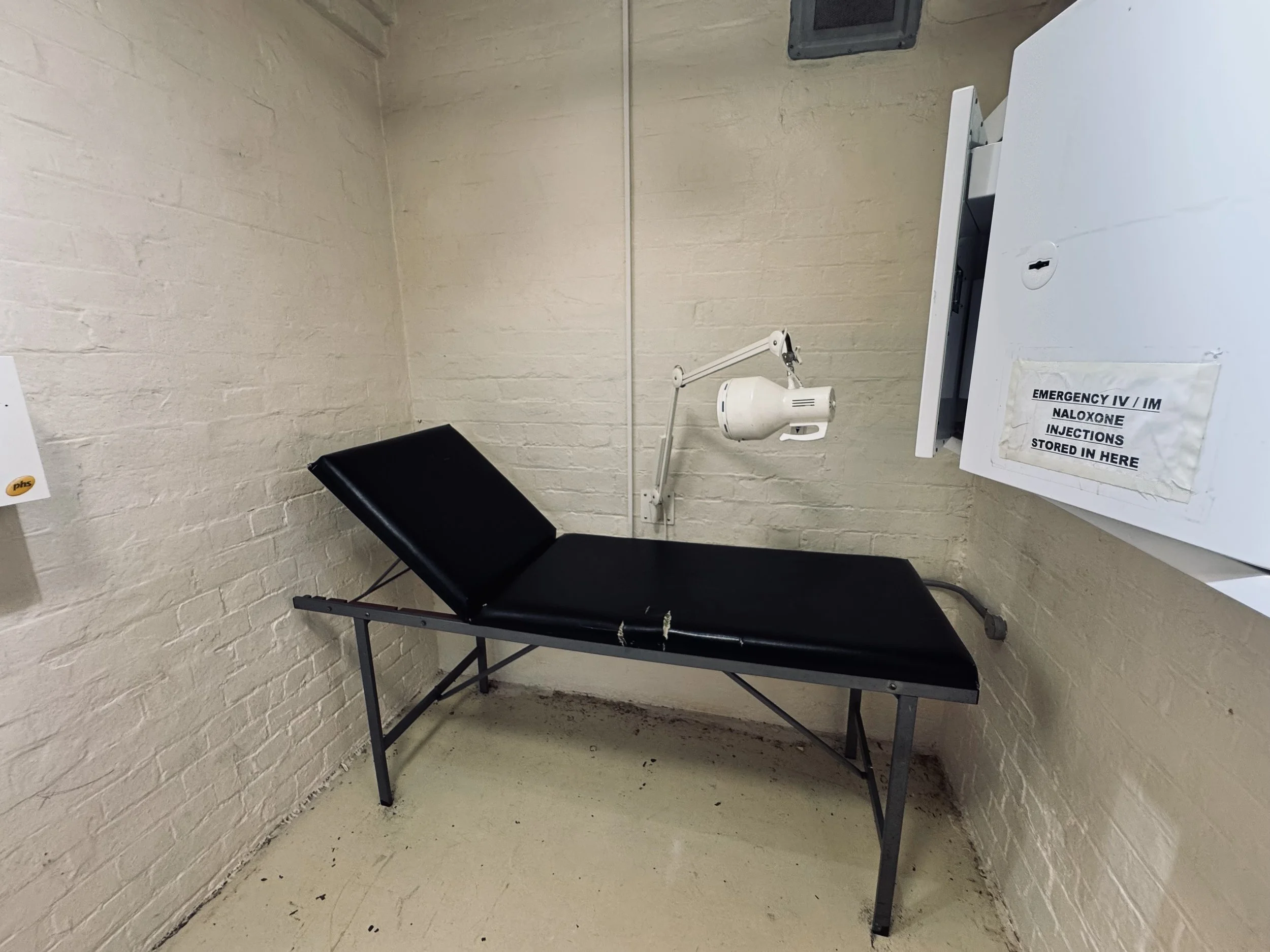 A small, empty room with white brick walls, containing a black padded examination table, a wall-mounted dental or medical light, and a white cabinet labeled for naloxone injections.