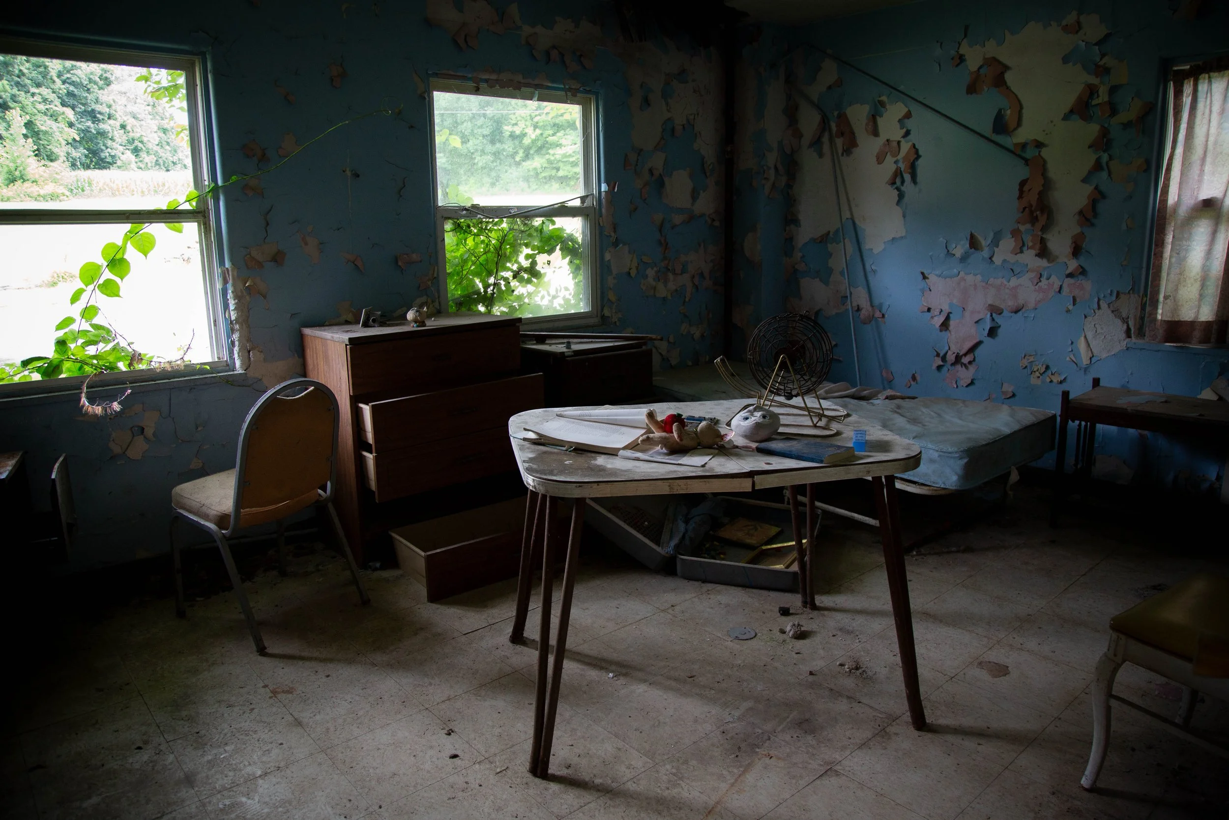 An old, empty room with a broken window, a white chair draped with a polka-dotted cloth, and a dirty mattress on the floor. The room is dusty and in disrepair.