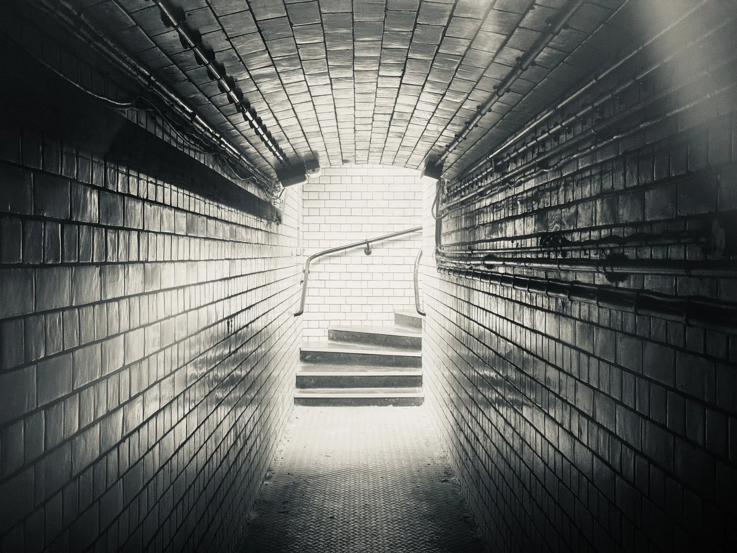 Dark underground stairwell with brick walls, metal railings, and steps lit by bright light at the top of the stairs.