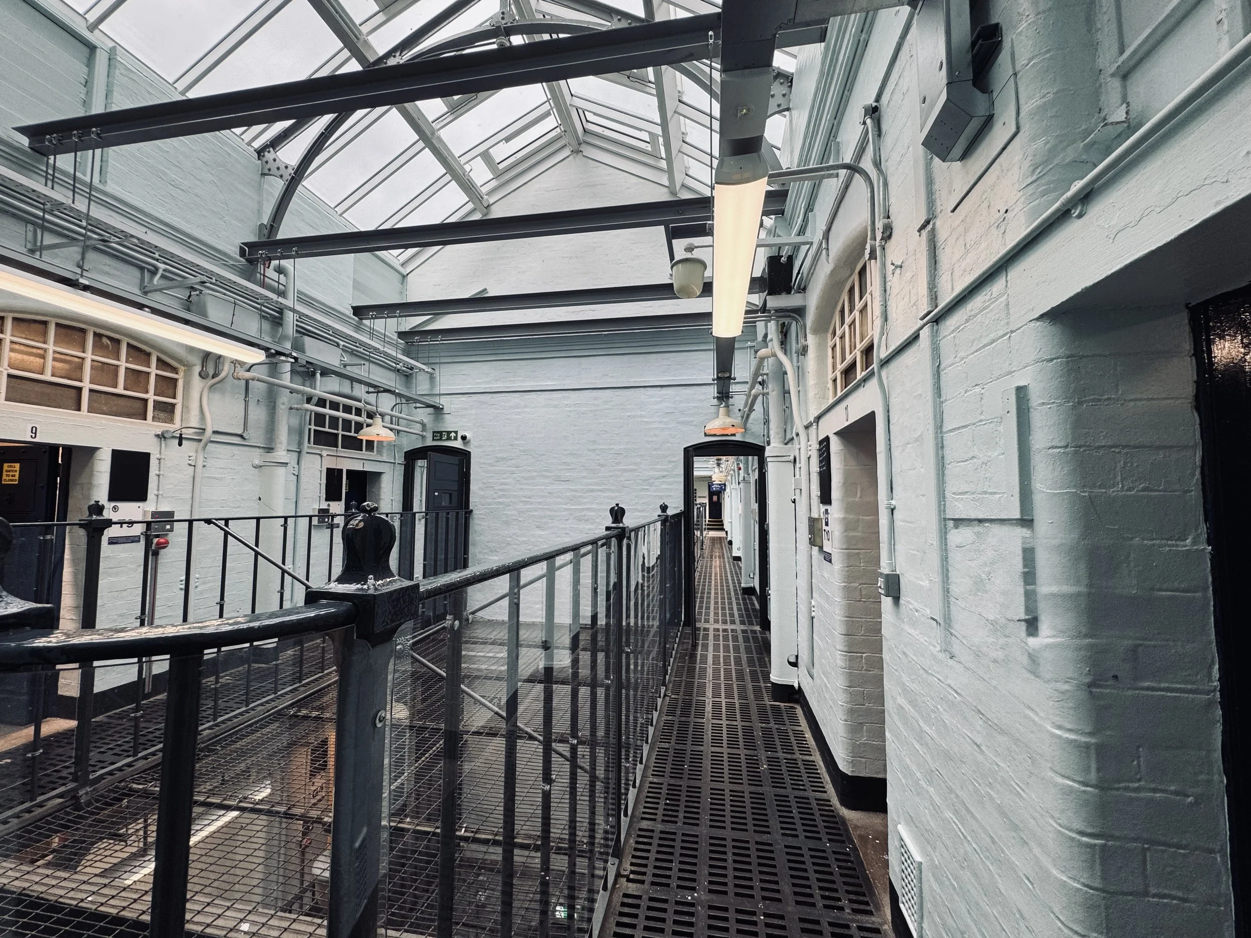 Indoor hallway with metal grated flooring, white brick walls, black metal railing, and a glass skylight ceiling with exposed beams.
