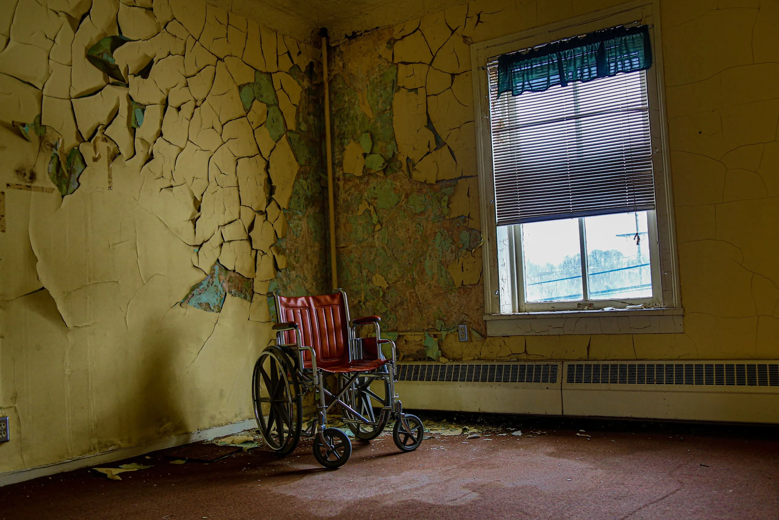 A black and white photo of an empty wheelchair in a dark corridor with closed doors on both sides and pipes on the ceiling.