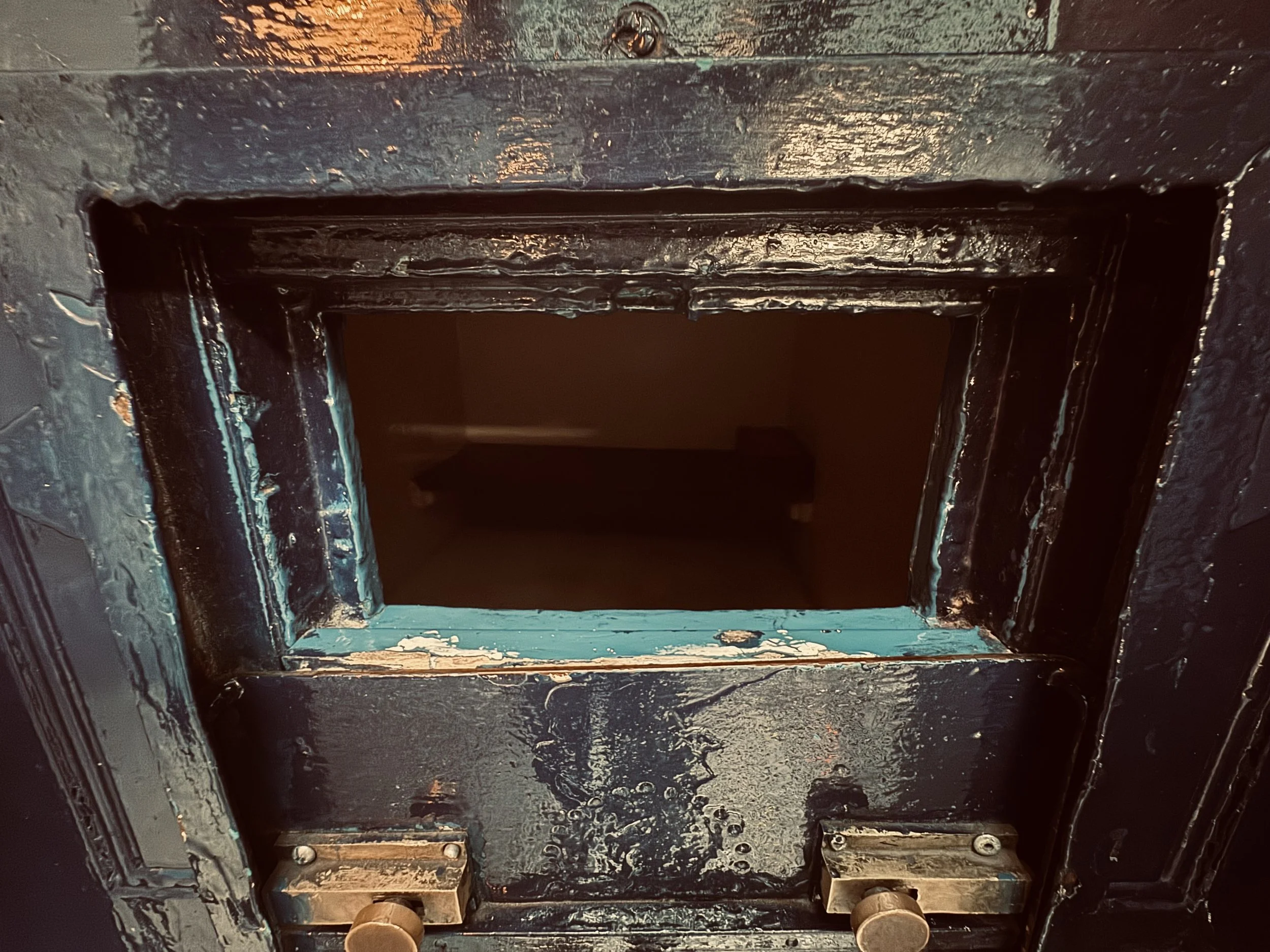 Close-up of a rectangular opening in a black, textured surface with visible bolts and chipping paint, revealing a dark interior space.