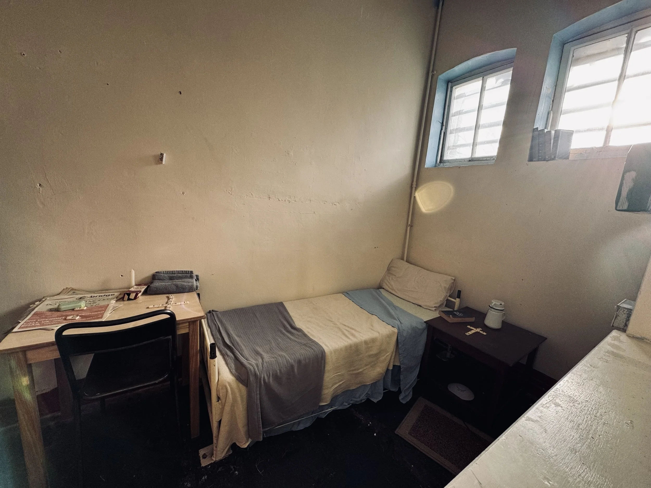 A small, modest bedroom with a twin bed covered in a mix of beige, gray, and blue sheets, placed against a beige wall with two small windows. There is a wooden nightstand with a lamp, a remote, and some crosses. A wooden desk with papers, glasses, and a towel is next to a black chair. A white textured surface is visible in the foreground.