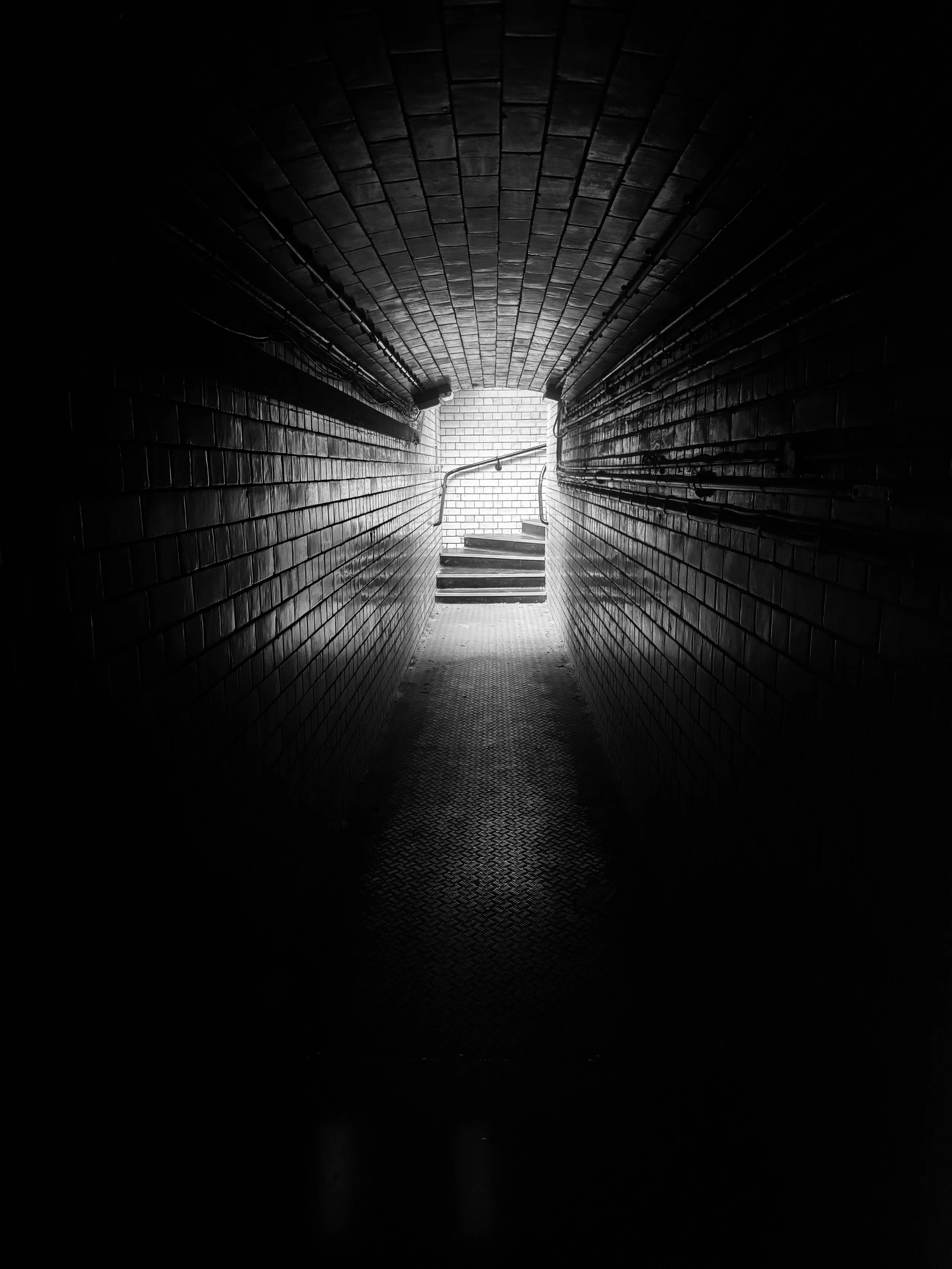 Dark underground tunnel with brick walls and a bright exit at the end, stairs leading up to a brighter area, metal handrails along the walls.