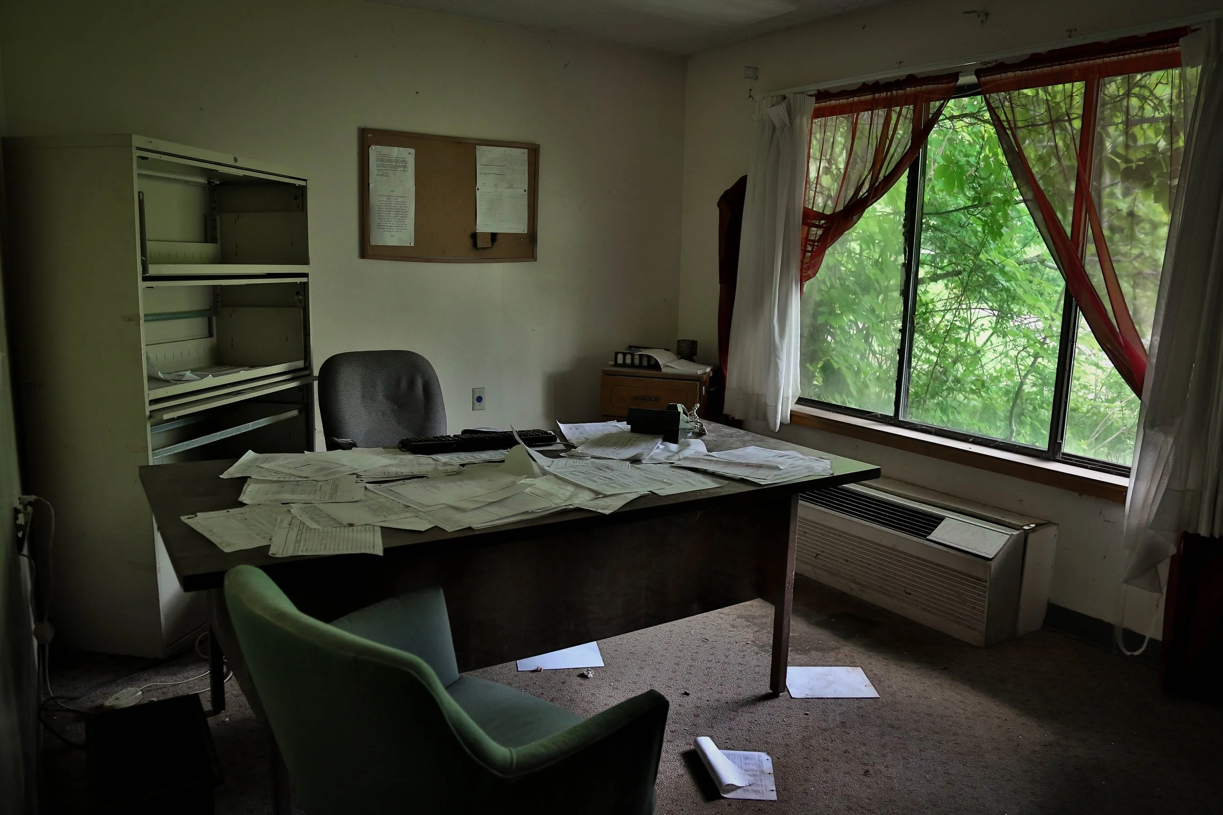 An office with a cluttered desk, paper documents, a black office chair, and an empty filing cabinet. The room has a large window with curtains, an air conditioning unit, and a bulletin board on the wall.