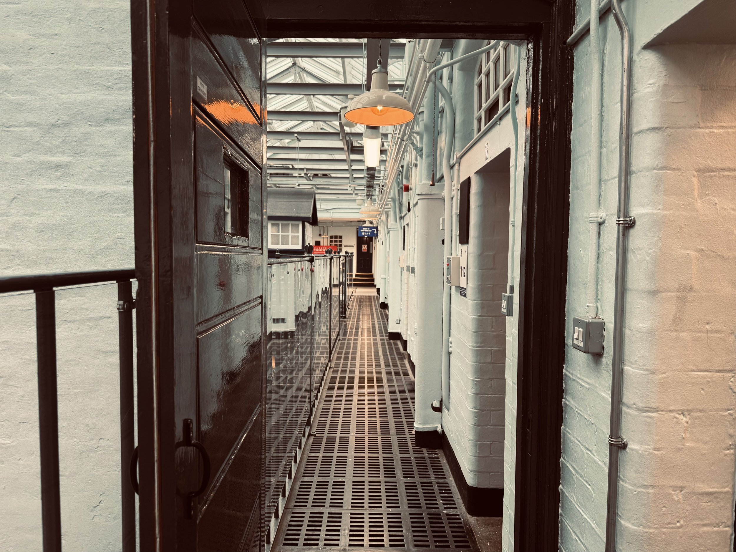 Industrial-style hallway with white brick walls, black metal flooring, overhead industrial pendant lights, and various utility pipes and boxes along the walls. The hallway leads to a door at the end.