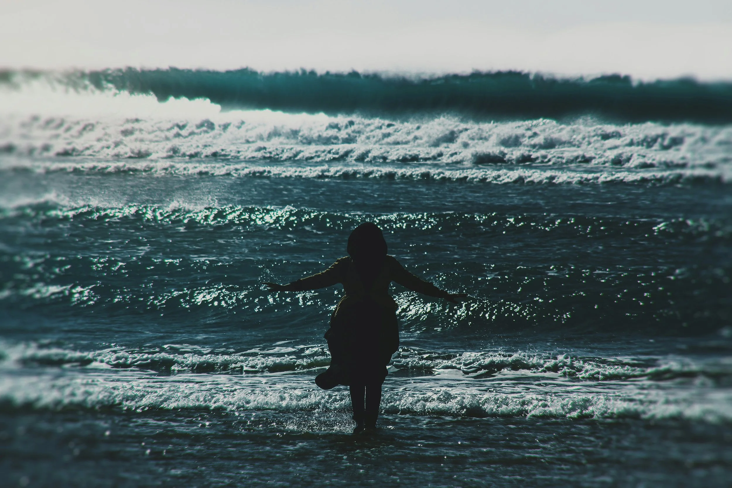 Silhouette of a woman standing at the ocean's shore with arms outstretched, waves crashing in the background, ready to accept the healing power of saltwater.
