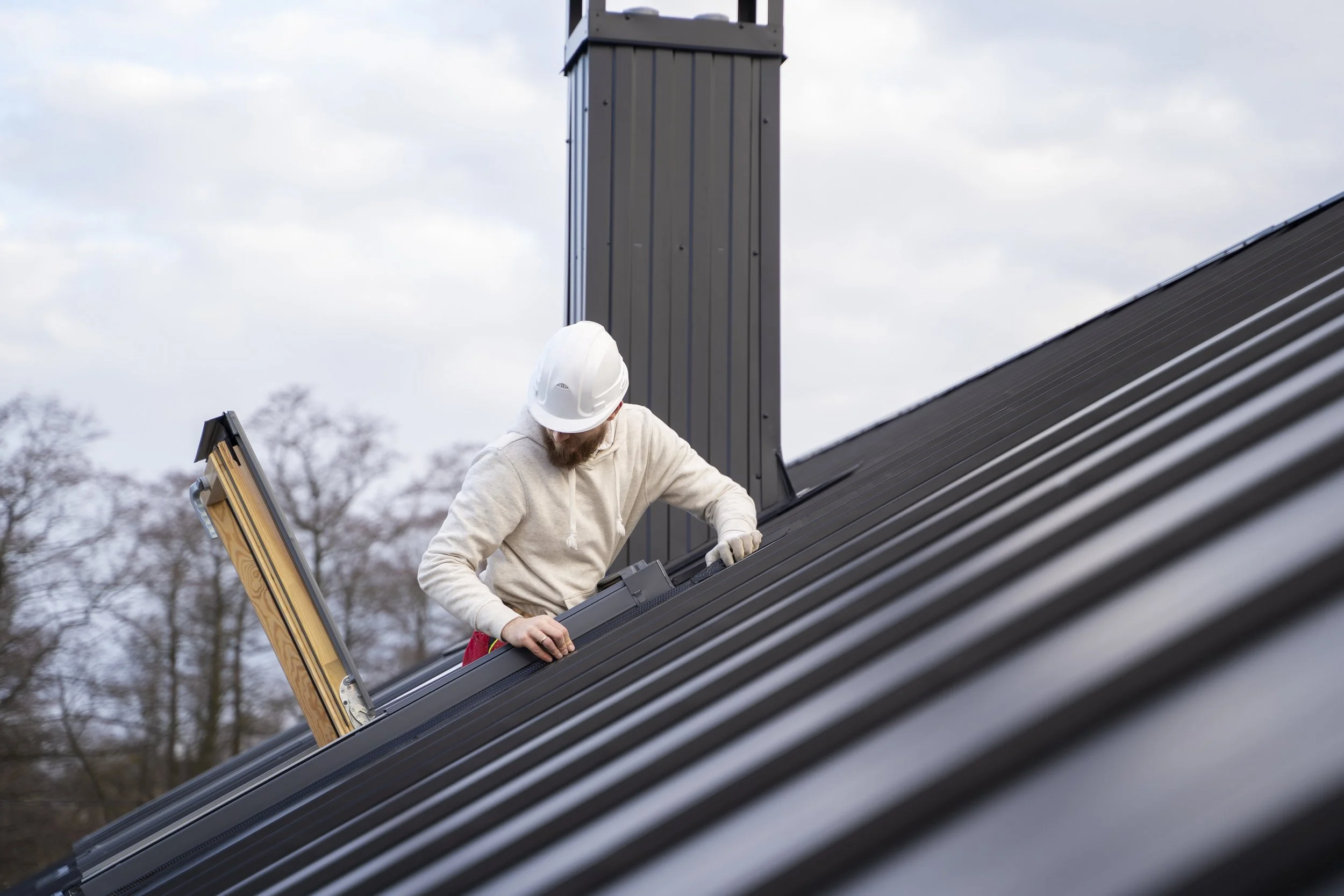 A construction worker wearing a white helmet and beige hoodie installing metal roofing panels on a sloped roof outside.