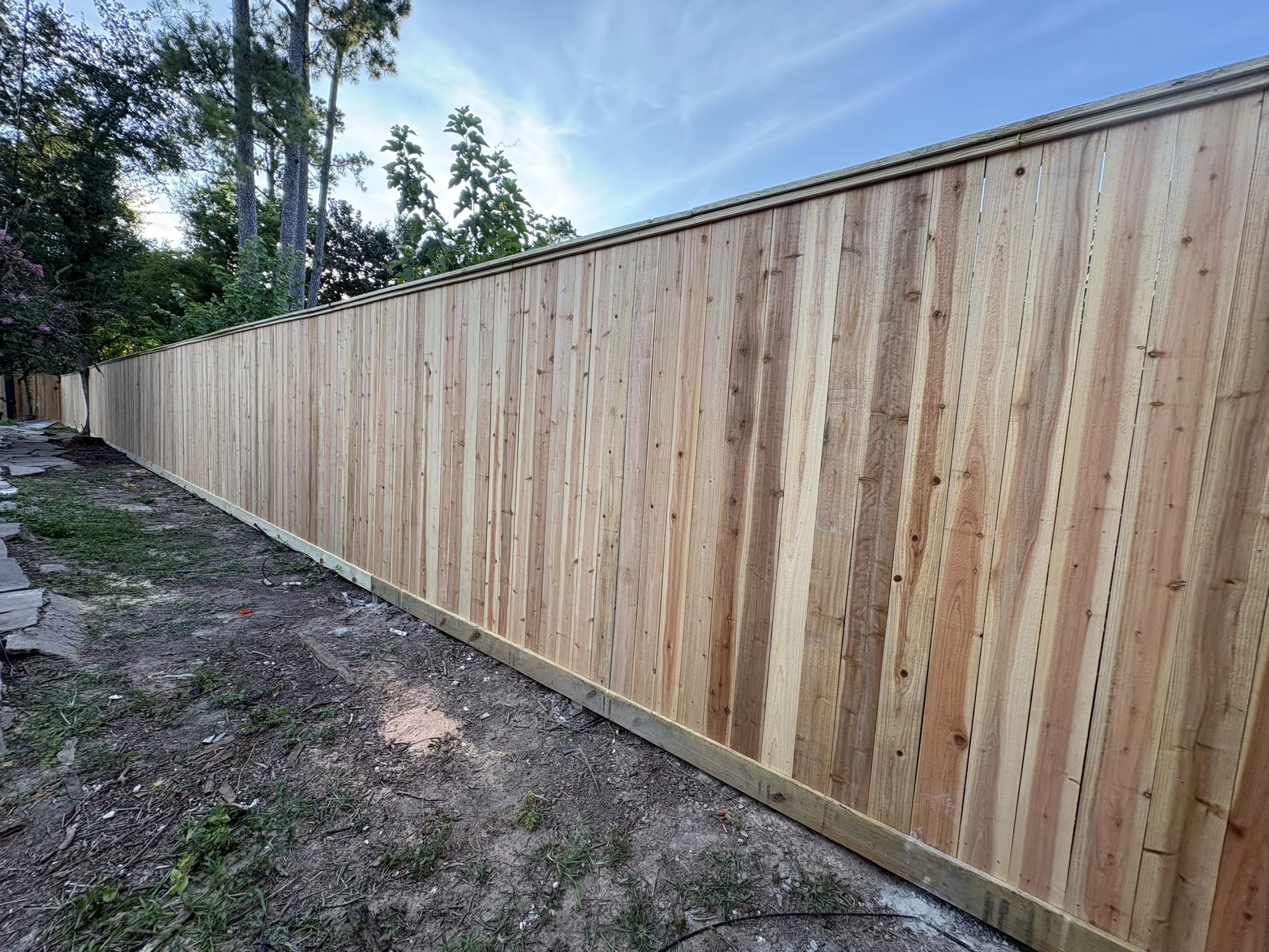 A newly constructed wooden privacy fence with vertical planks and a sloped top, situated along a dirt and grass area, with trees and a blue sky in the background.