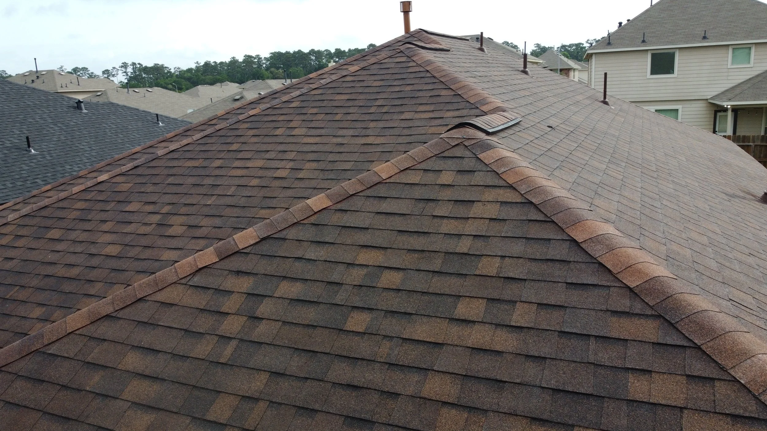 A view of rooftops with various colored shingles, including brown, gray, and black, on residential houses in a neighborhood with trees in the background.