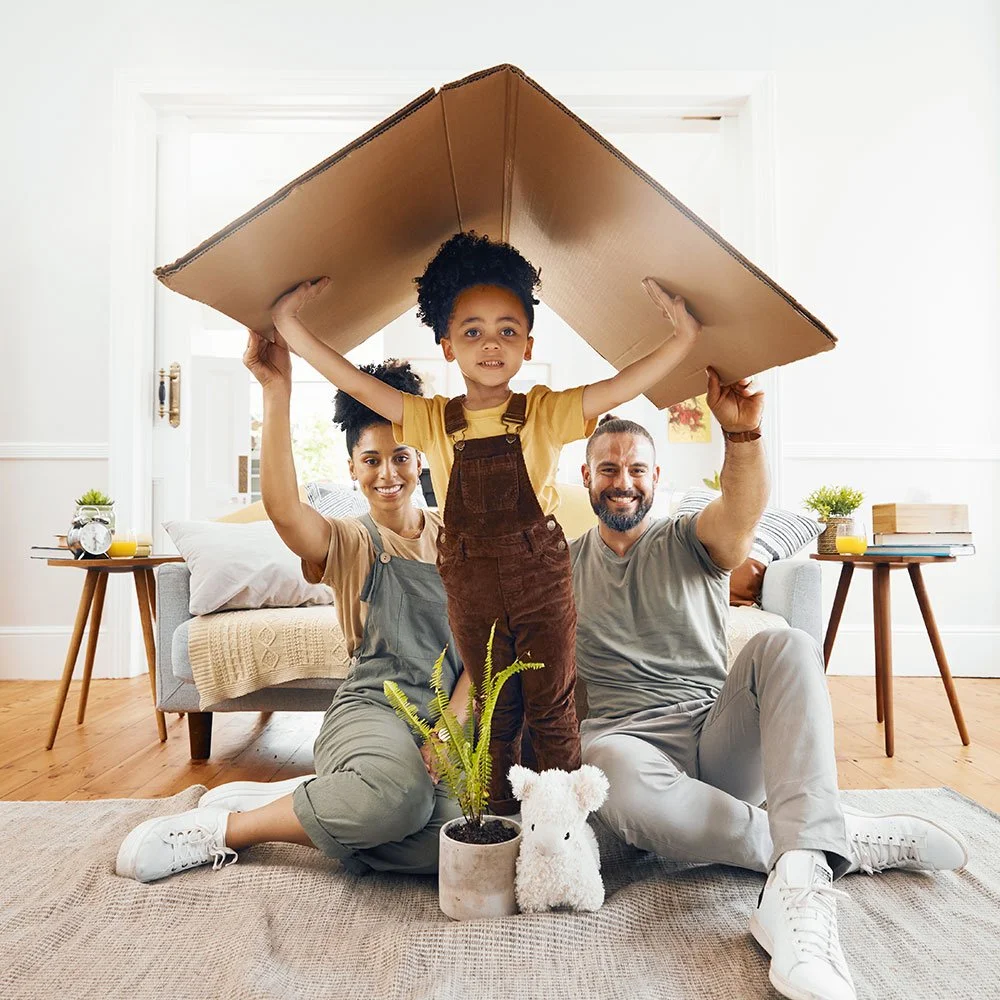 Three people, a girl standing on a cushion with a large cardboard box over her head, and two adults sitting on the floor behind her, smiling and supporting her. They are in a bright living room with a couch, side tables with small plants and books, and a window in the background.