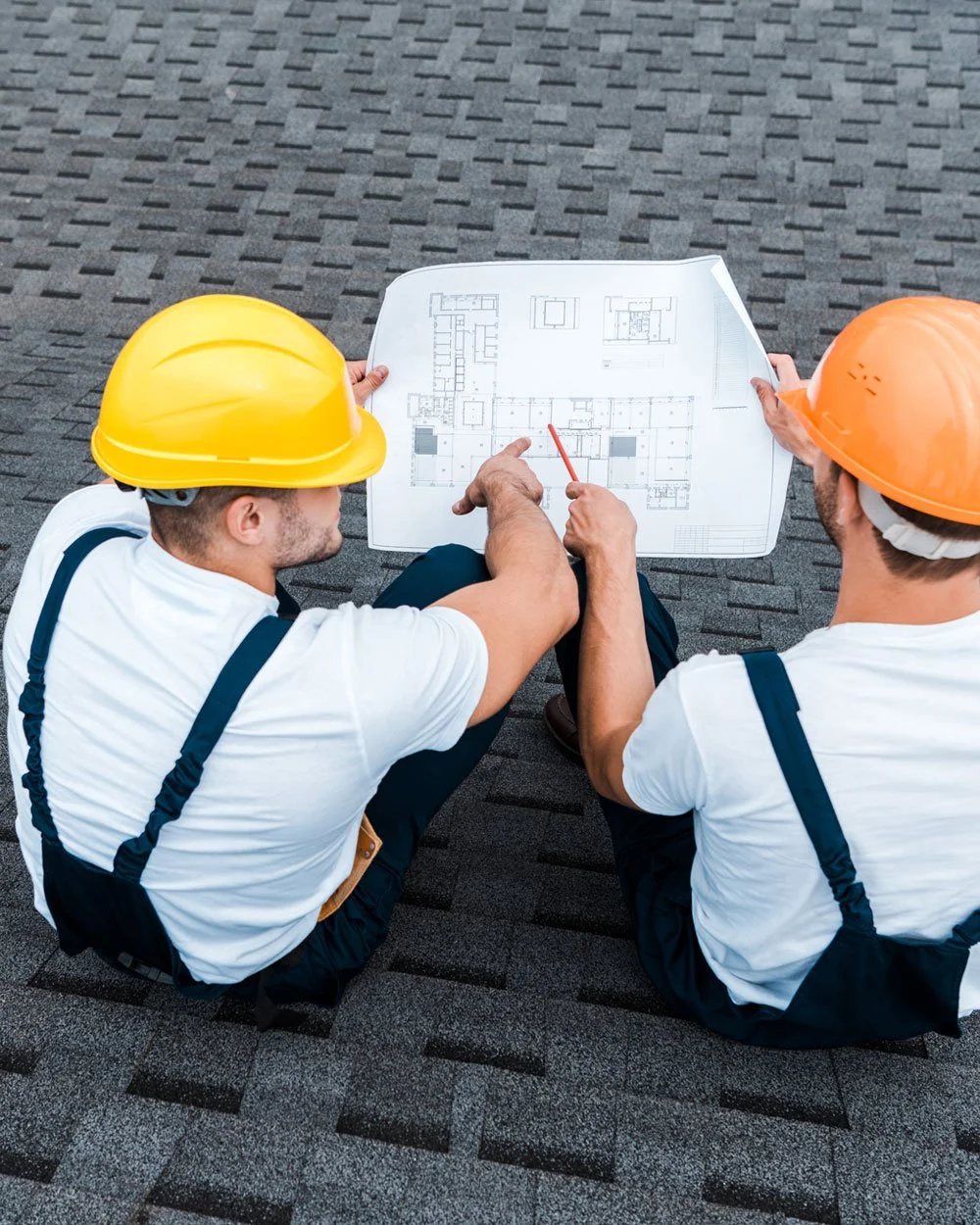 Two construction workers with yellow and orange helmets sitting on roofing shingles discussing blueprints.