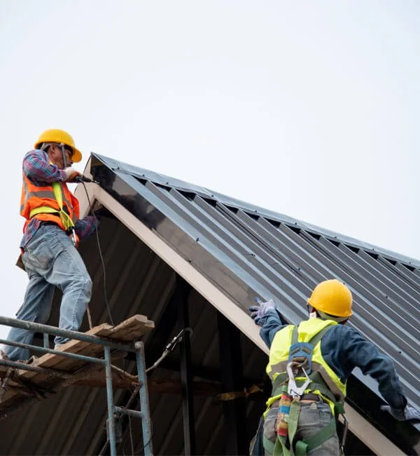 Two construction workers wearing safety helmets and vests working on the roof of a building during daytime.