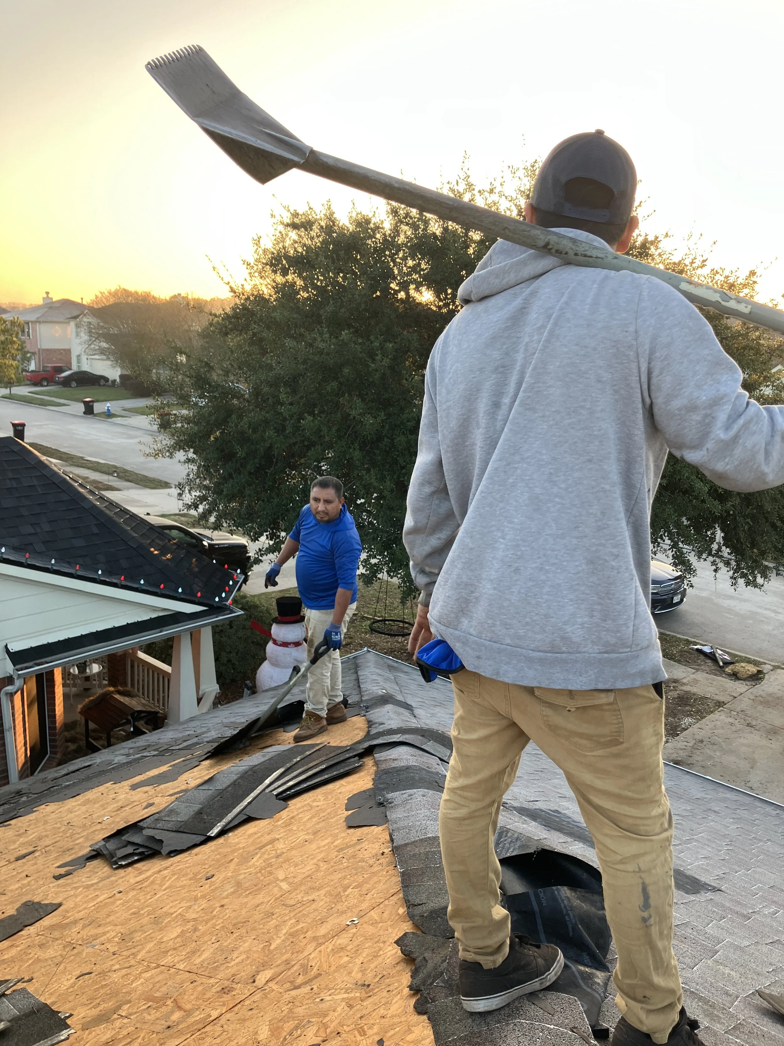 People working on a roof during sunset, with one man holding a rake over his shoulder, and another man in a blue shirt and gloves nearby.