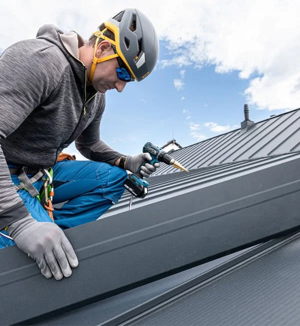 A person wearing a helmet, gloves, and outdoor clothing installing or repairing a metal roof with a power drill.