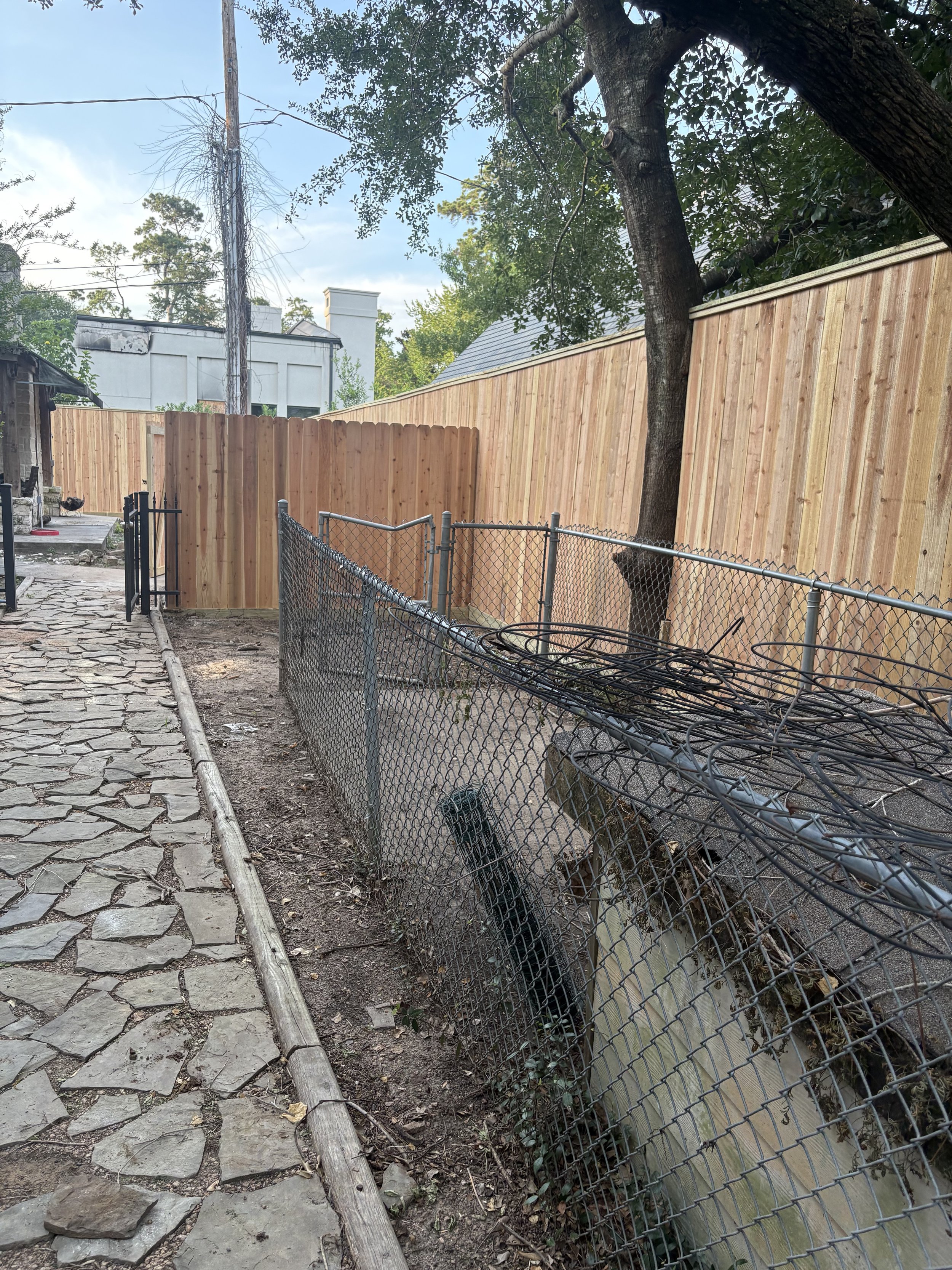 A backyard with a wooden fence, a tree, a chain-link fence, and a stone pathway. There is a utility pole with wires and some houses in the background.