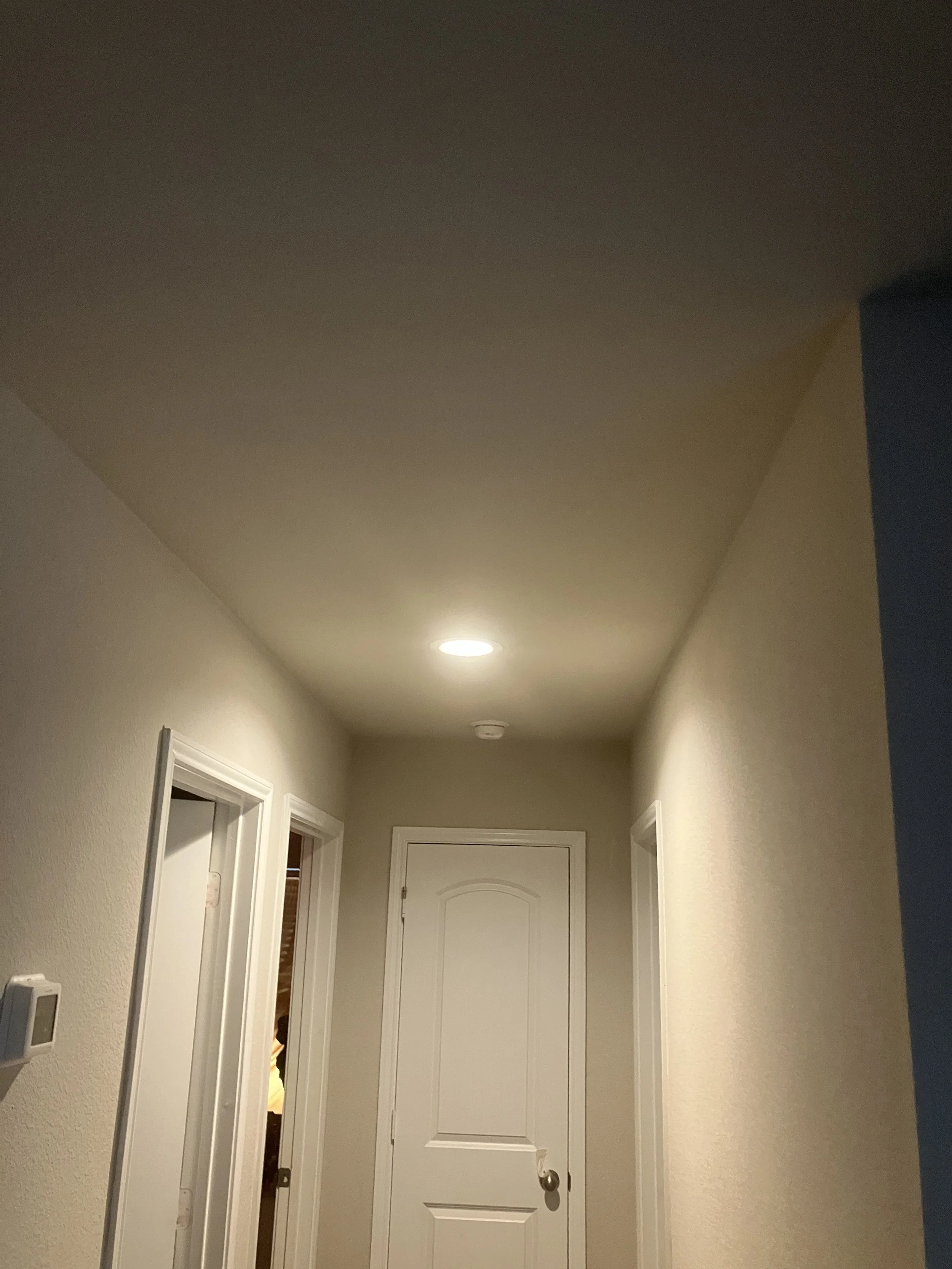 A hallway with beige walls, white trim, and a closed white door at the end. There are two doorways on the left side of the hallway and a ceiling light fixture.