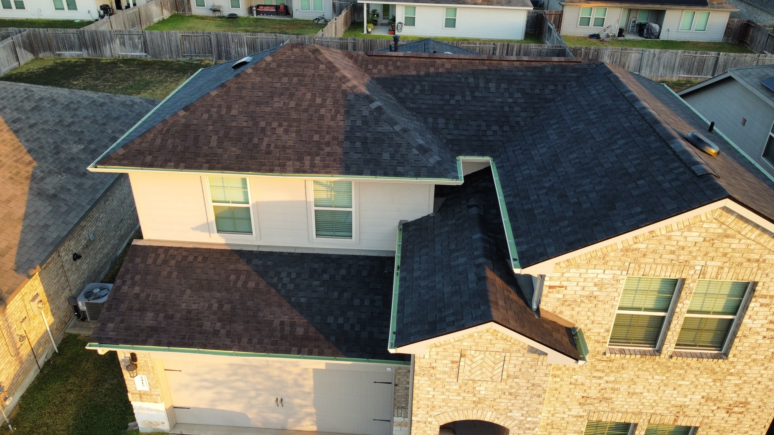 An aerial view of a house with a multi-section roof, showing partial asphalt shingle roofing in different colors, surrounded by neighboring houses and fenced yards.