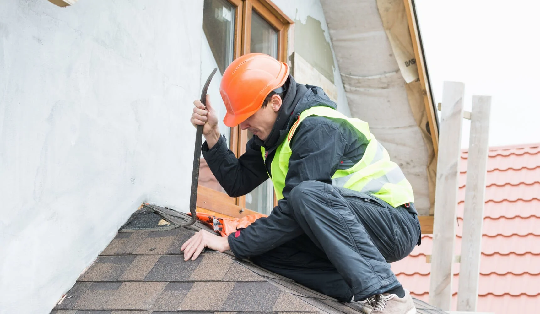 A construction worker wearing an orange safety helmet and a reflective yellow safety vest crouching on a roof, examining or working on shingles near a window.